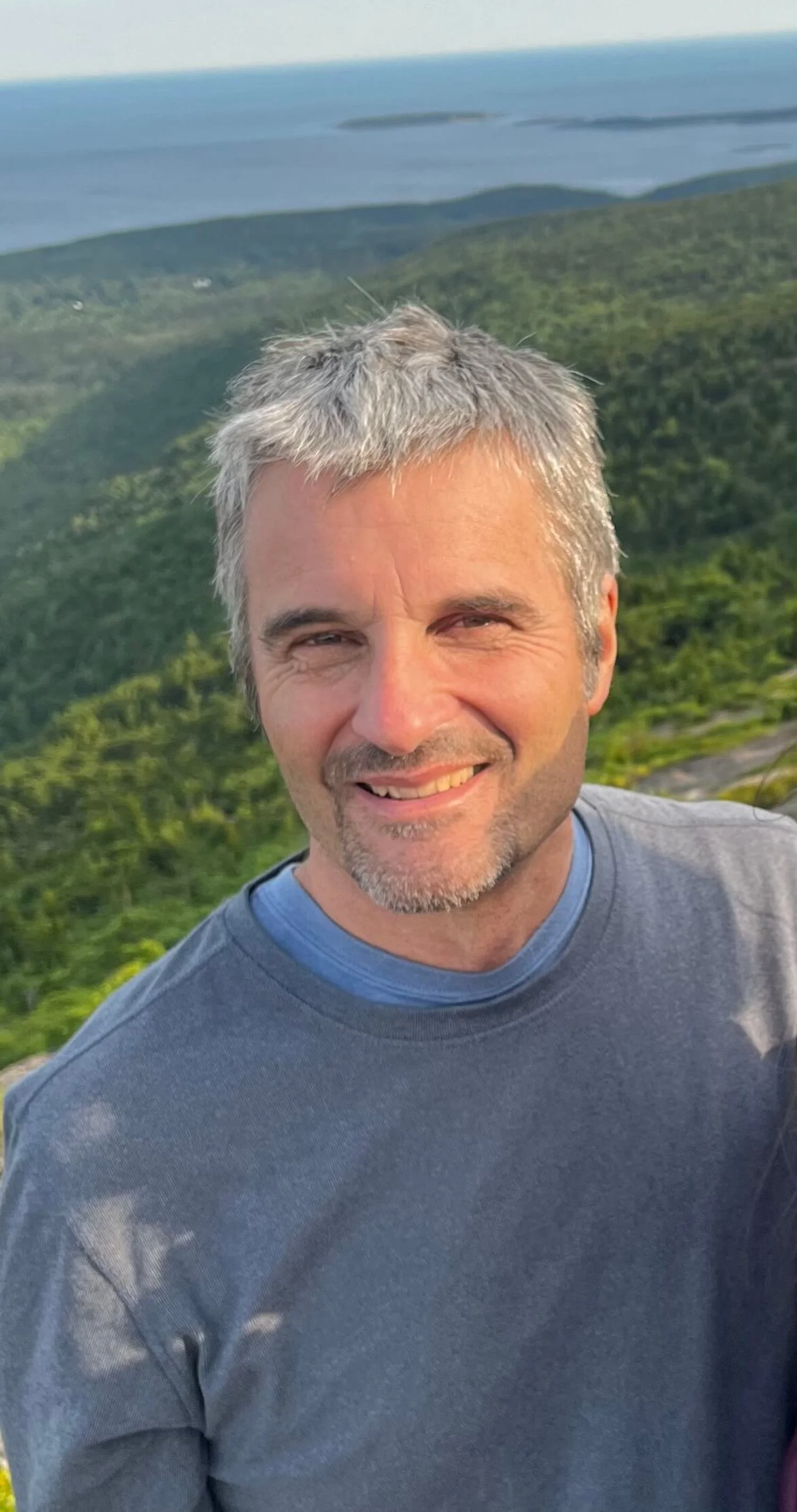 John Monks, founder of Vermont Tree Goods is smiling with salt-and-pepper hair and a beard, wearing a blue shirt, standing on top of a mountain at Acadia National Park