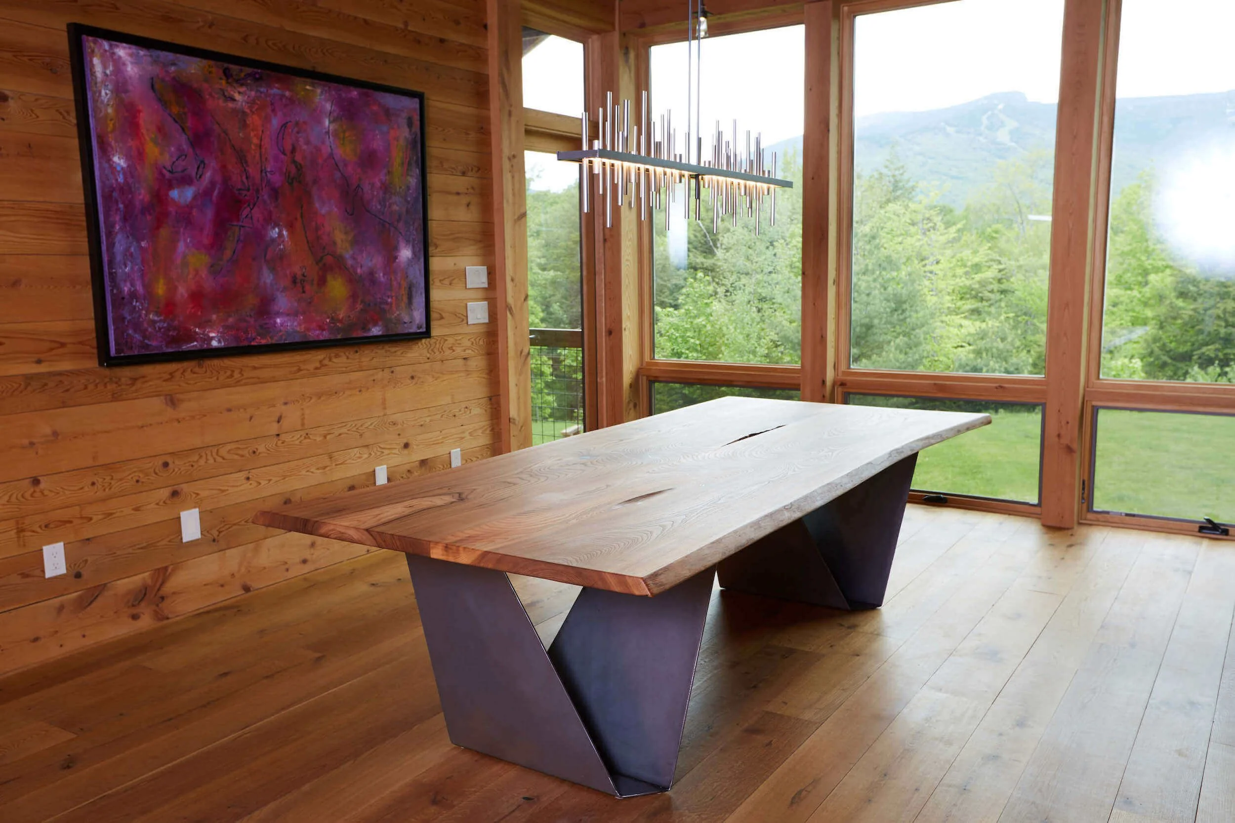 A black walnut live edge dining table with a metal base inside a room with large glass windows looking out at a Vermont ski mountain.