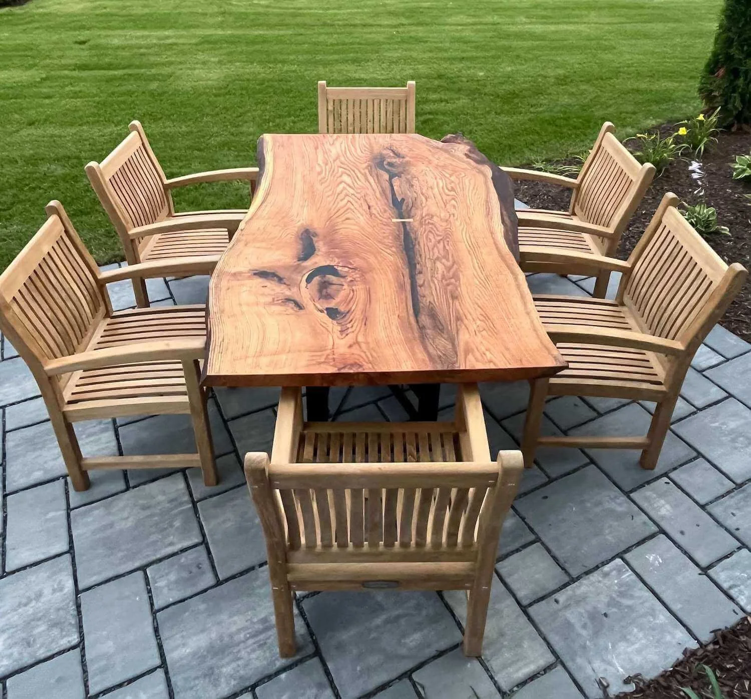 Beautiful oak table with natural edges and dark swirling grain on an outdoor patio table with six wooden chairs surrounding it, set on a stone-paved surface with a grassy lawn and garden bed with flowers in the background.