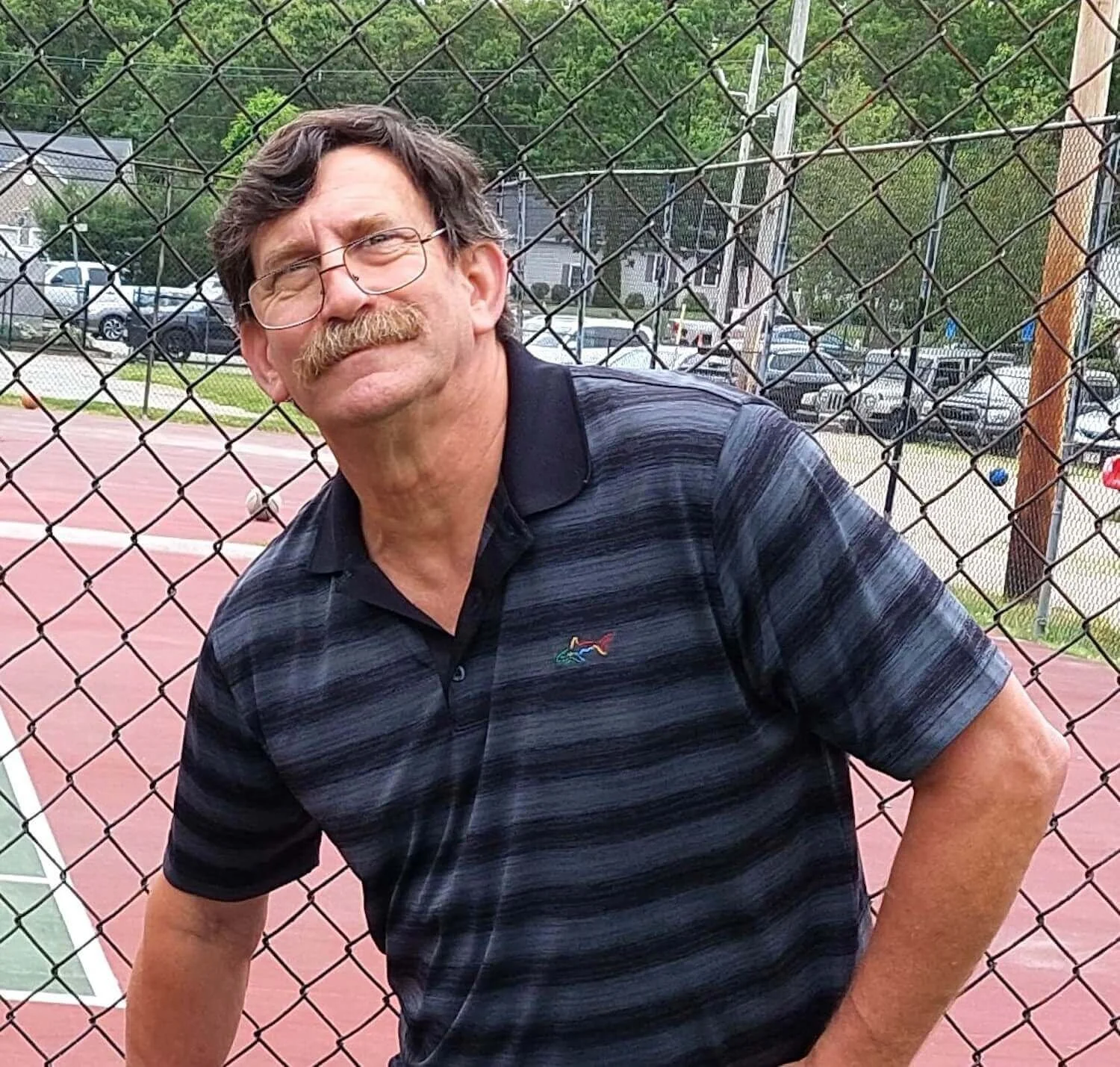 Dave Schnitzel, the lead operations worker at vtg with glasses and a moustache leaning against a chain-link fence at a tennis court, smiling and wearing a dark striped polo shirt.