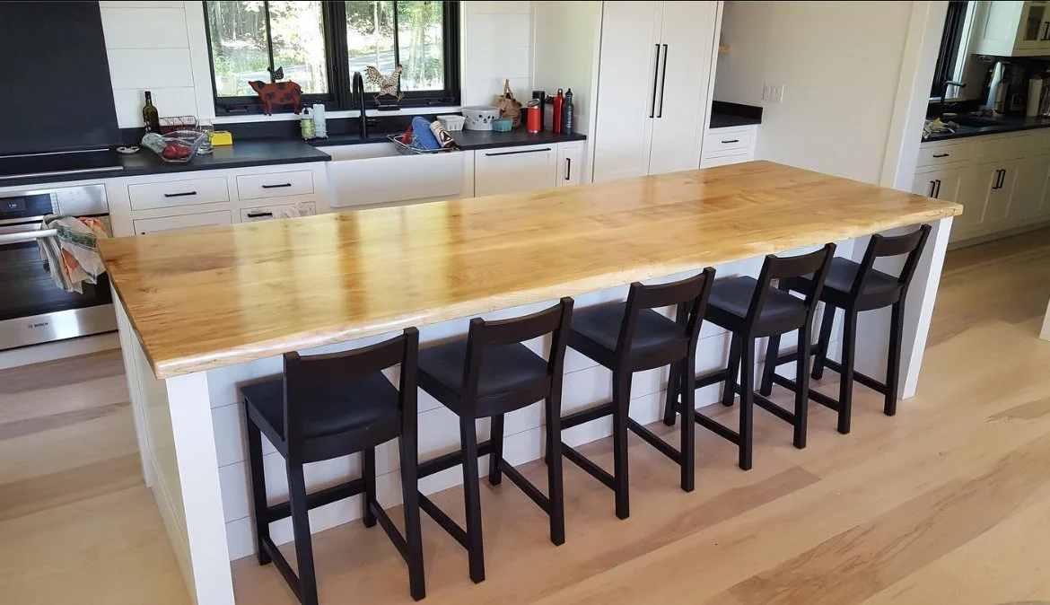 Extra-long live edge maple island countertop with natural bark edge, installed in a modern white kitchen with black bar stools — seats five comfortably