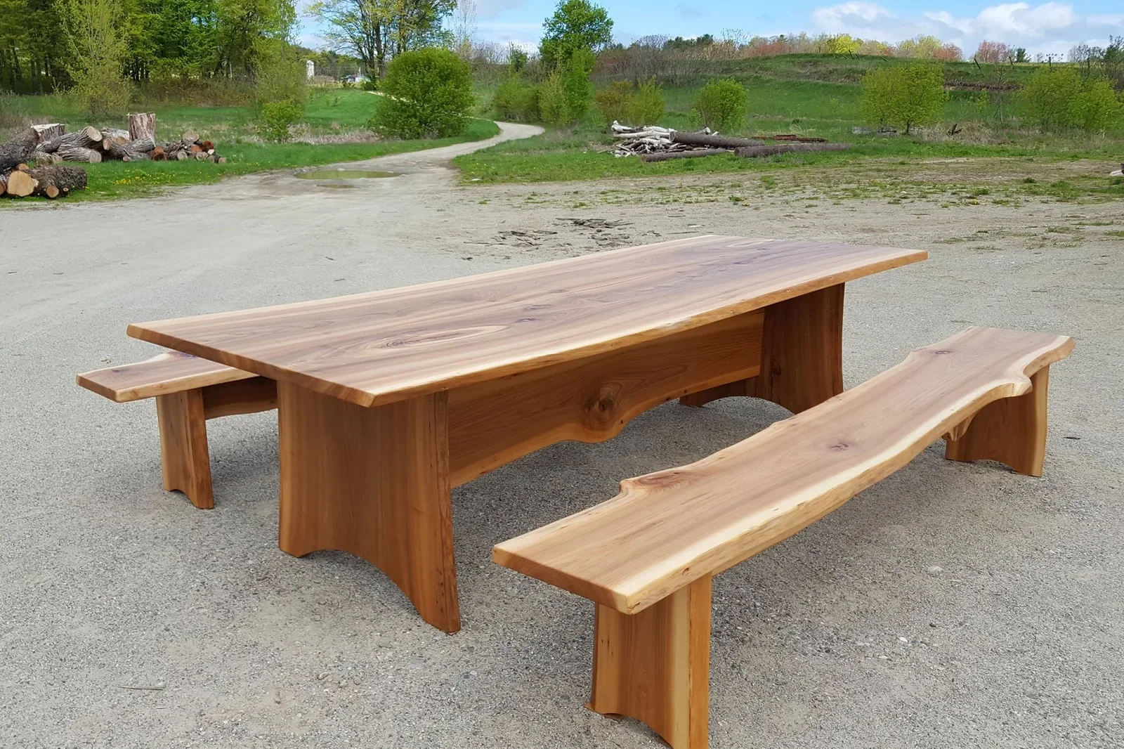 A black walnut meeting room table with two benches outdoors on a gravel surface, surrounded by green trees and a grassy landscape.