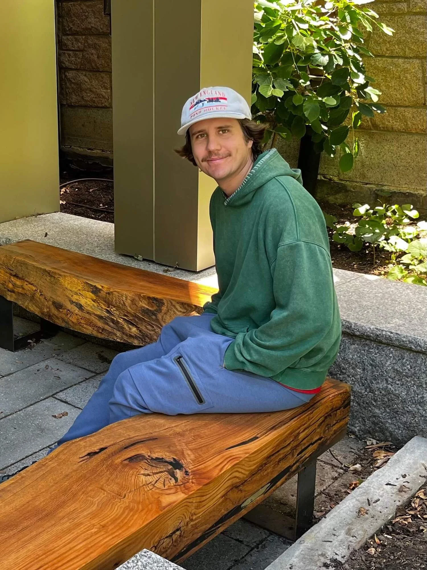 Carter Monks with brown hair, wearing a blue cap with red and white text, a green hoodie, and gray pants, sitting on a VTG wooden bench outside Harvard Divinity School.