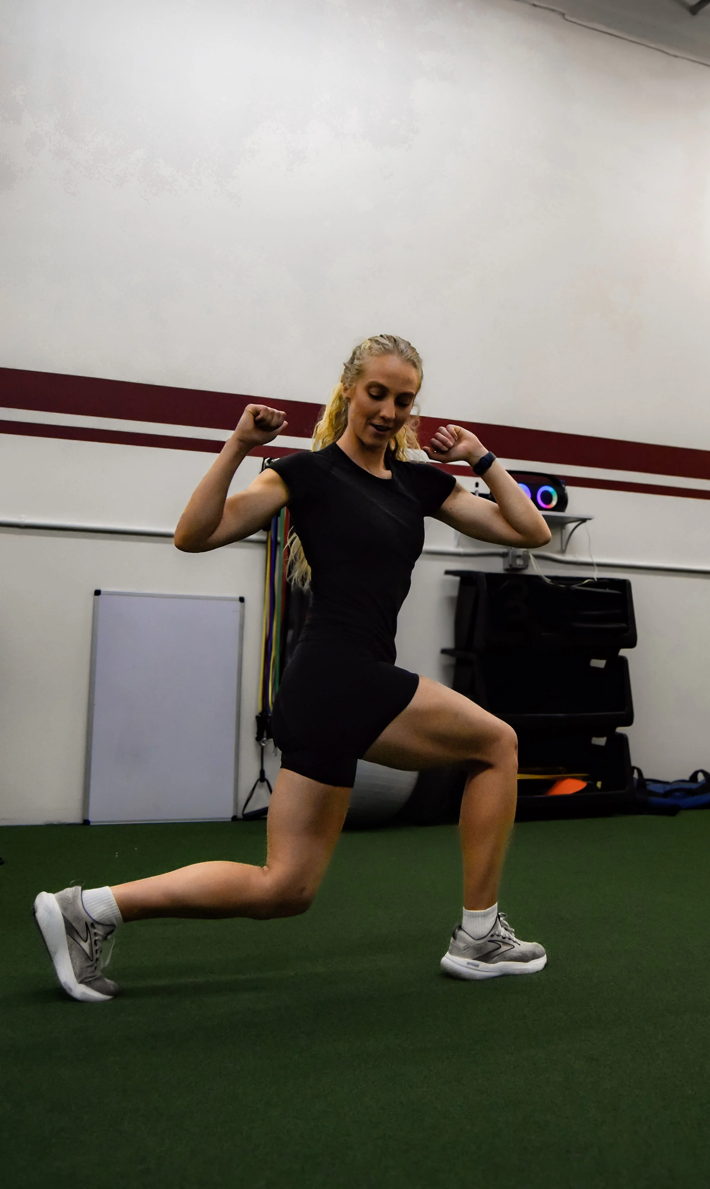 A young woman with blonde hair in a ponytail working out in a gym, performing a lunge exercise on green turf, with her arms bent and fists clenched, wearing a black T-shirt, black shorts, and gray sneakers.