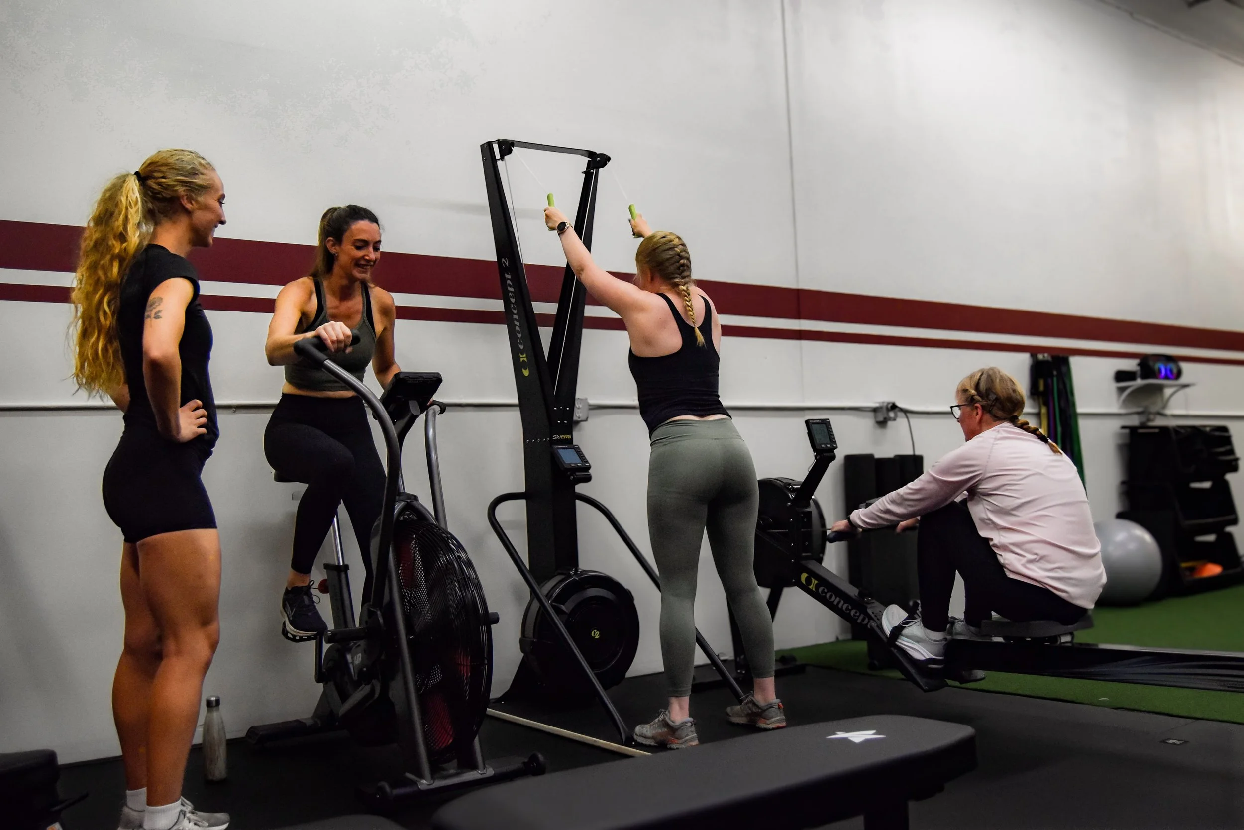 Two women in workout clothes high-five each other while doing a plank exercise in a gym with brick walls and large windows.