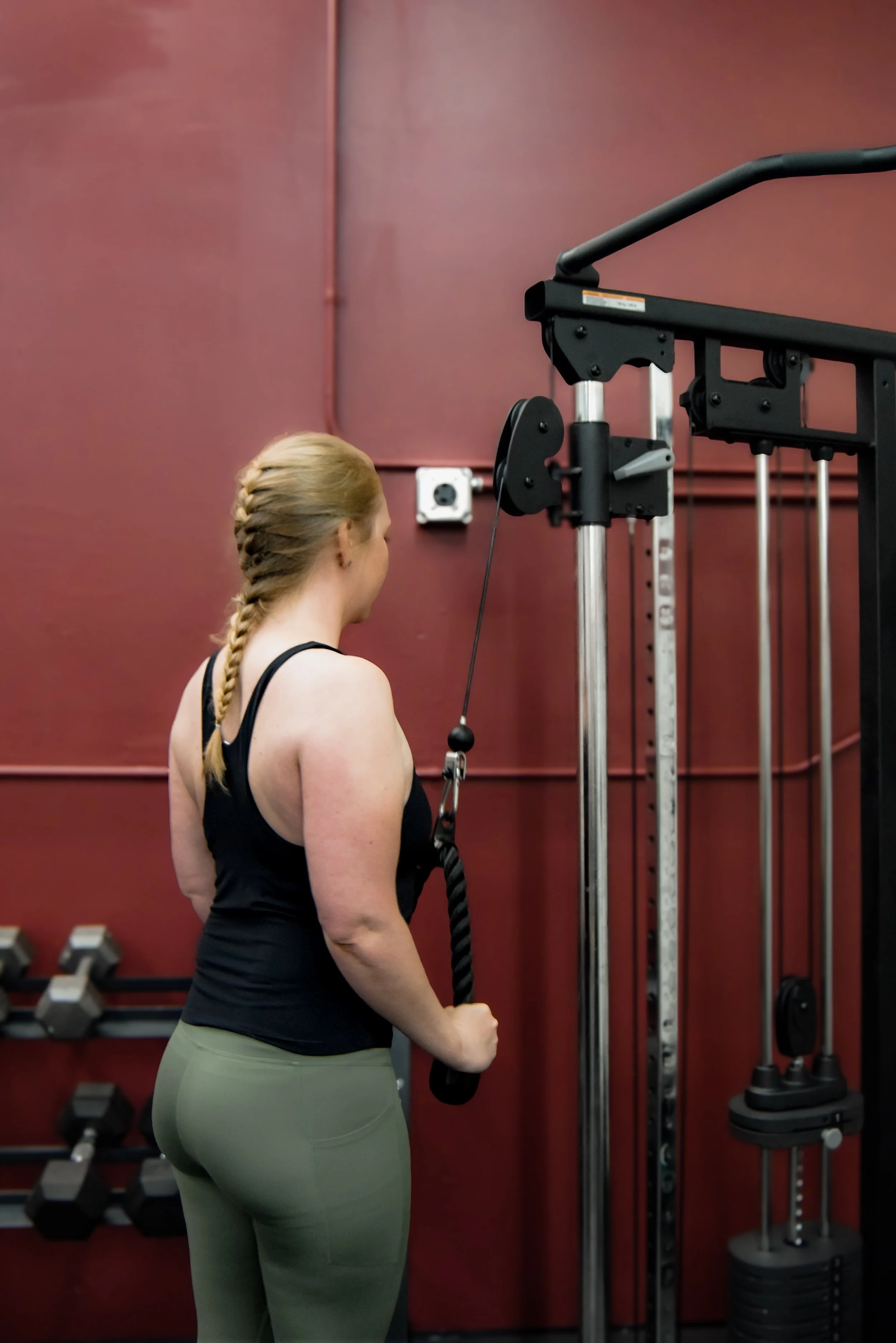 A woman with braided blonde hair wearing a black tank top and green leggings, standing in a gym, holding a cable handle attached to strength training equipment.