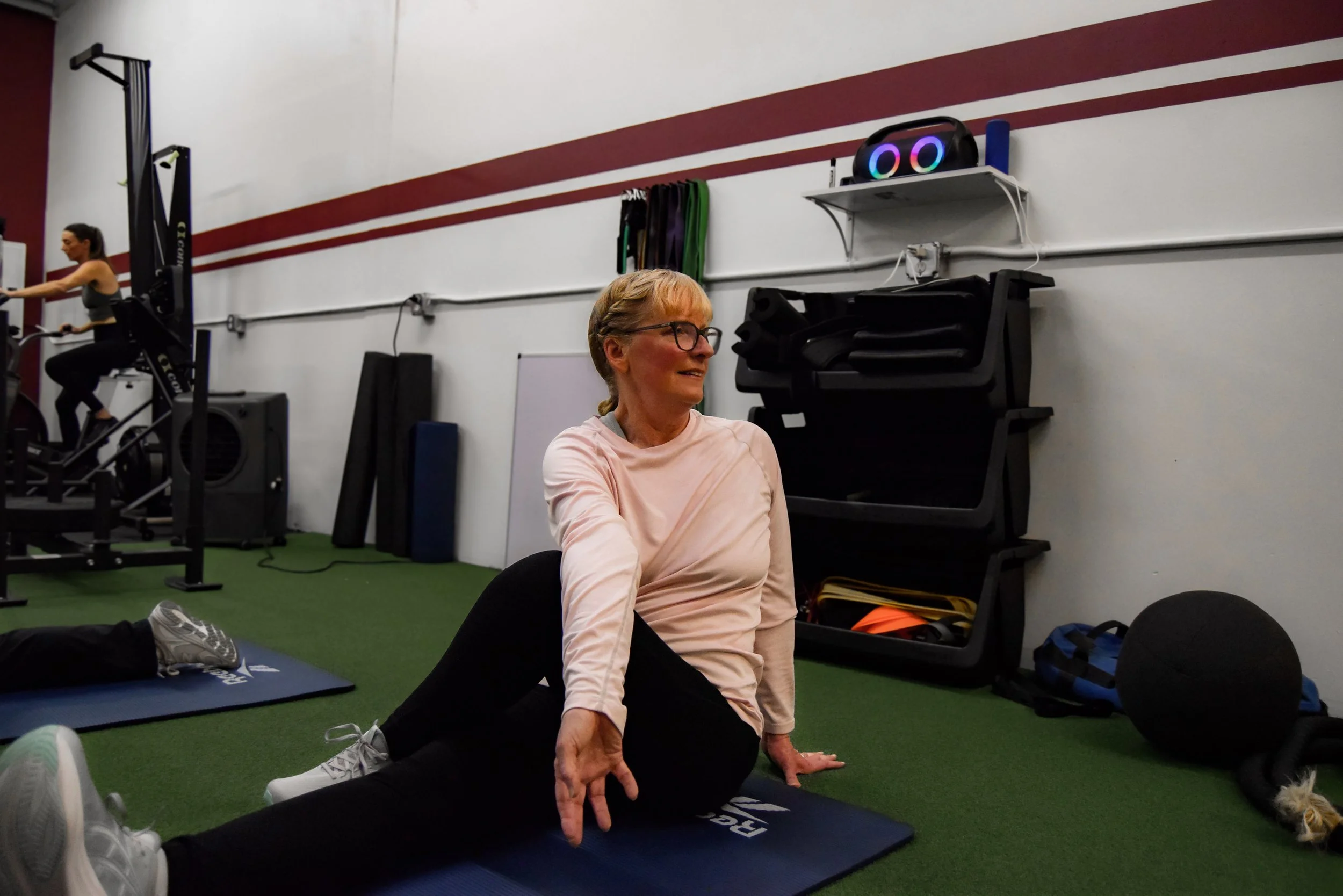 A woman performing a seated stretch on a yoga mat in a gym with fitness equipment and a man on an exercise machine in the background.