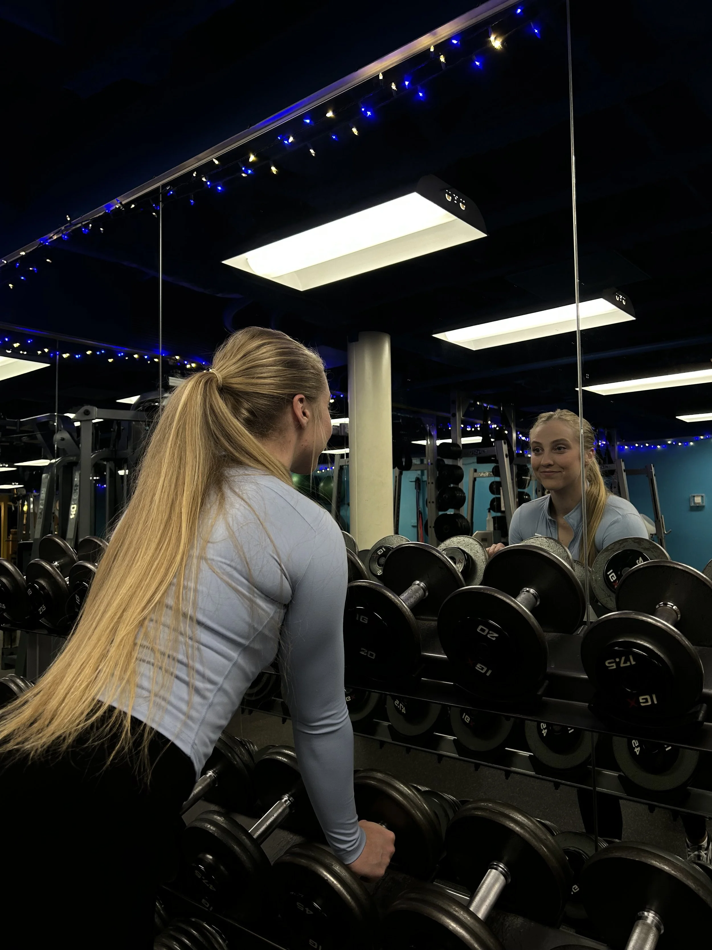 A woman with long blonde hair looking at herself in a gym mirror while surrounded by dumbbells, with blue accent lights and ceiling lights in the background.