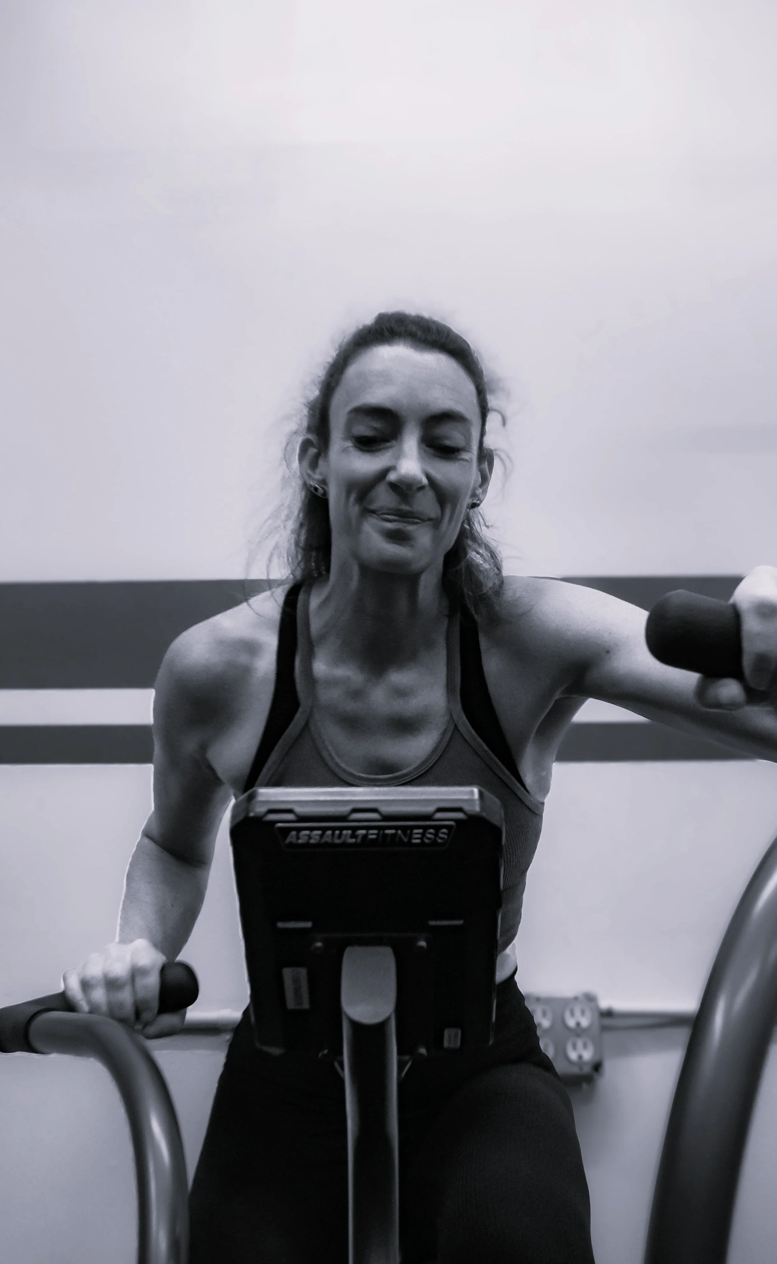 A woman working out on a stationary bike at the gym, wearing a tank top and smiling, with a wall and power outlets in the background.