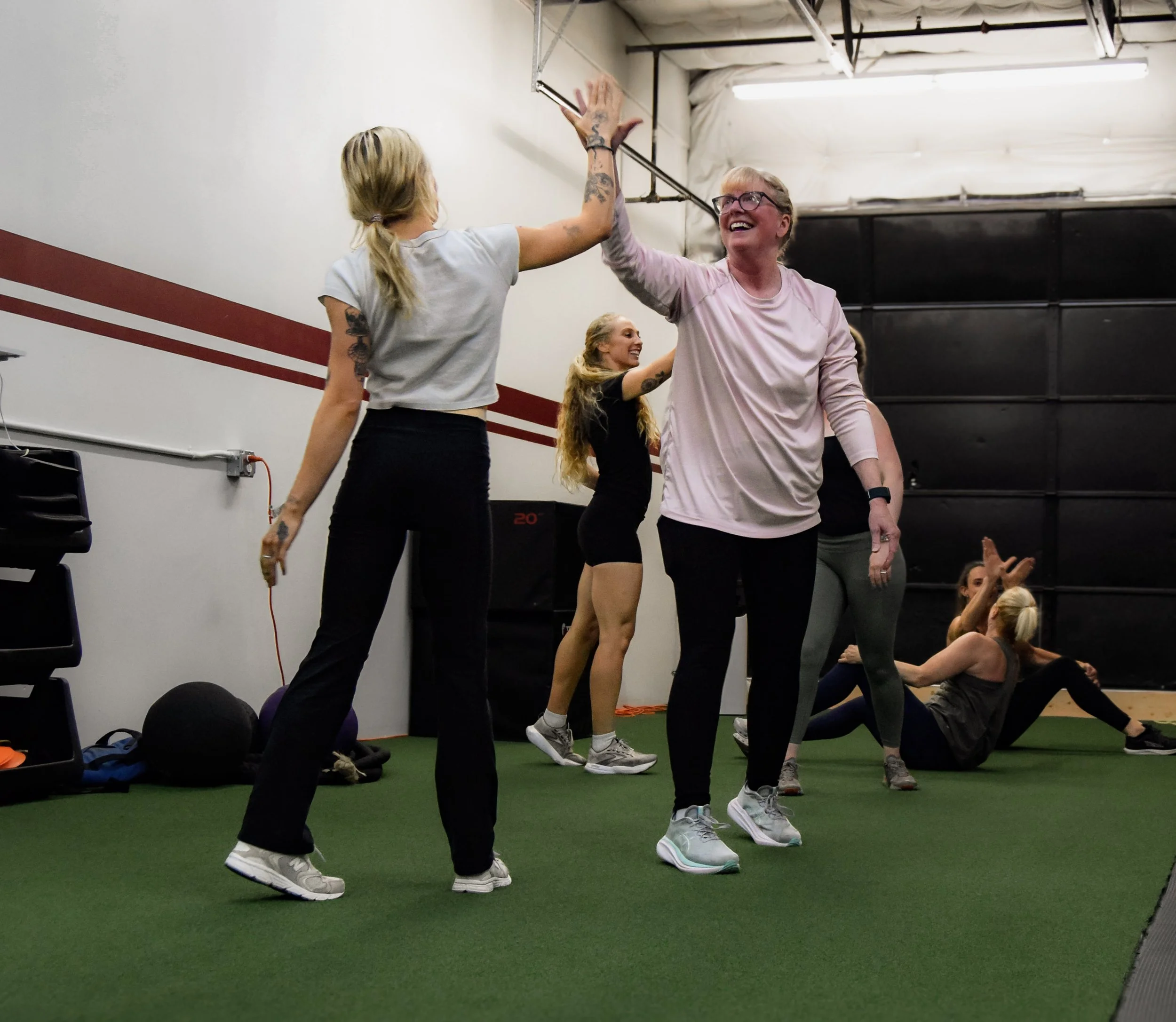 Group of people in a gym doing a workout, some high-fiving, with a woman in the foreground and a woman in the background smiling.