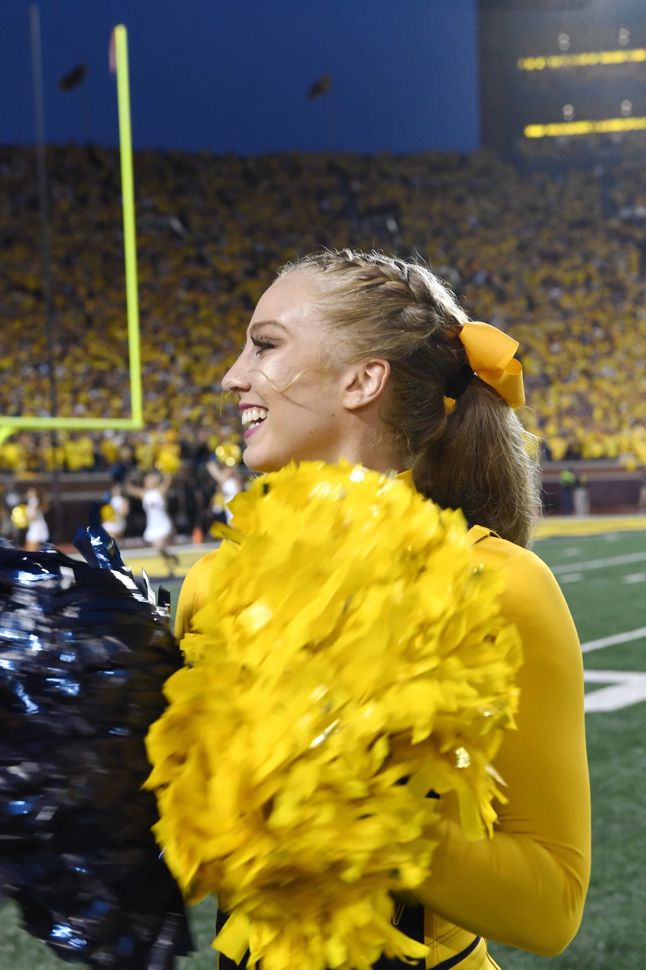 A smiling cheerleader in a yellow uniform and yellow bow in her hair, holding yellow and black pom-poms, standing on a football field during a game.