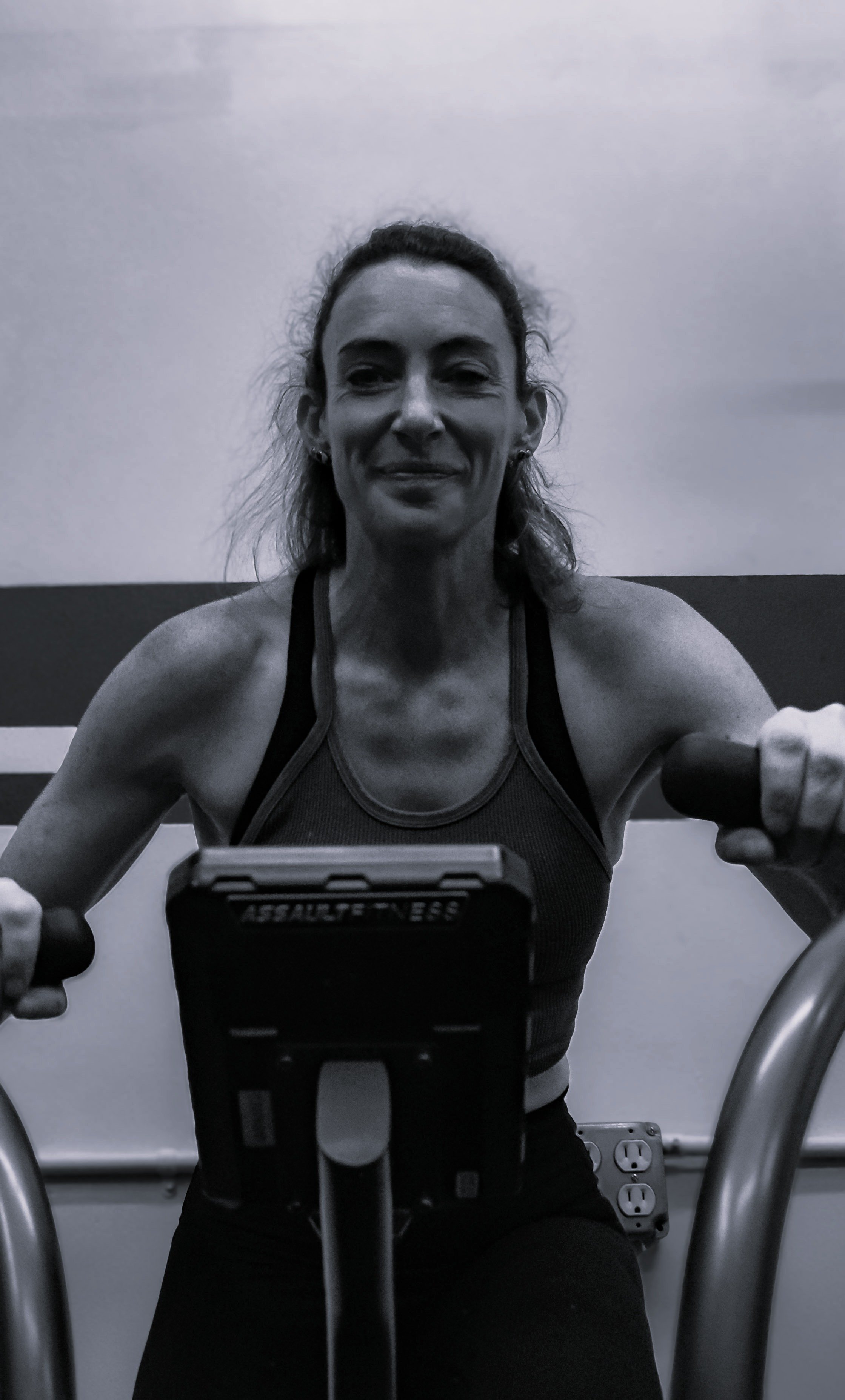 A woman at a gym taking a selfie on an exercise bike, smiling, wearing workout clothes, with gym equipment and outlets in the background.