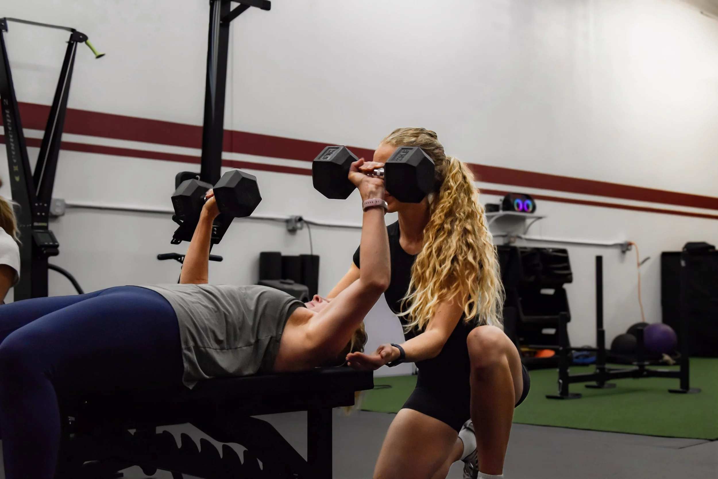Woman lifting dumbbells on an elevated platform inside a spacious gym with large windows, exercise machines, and a woman working out in the background.