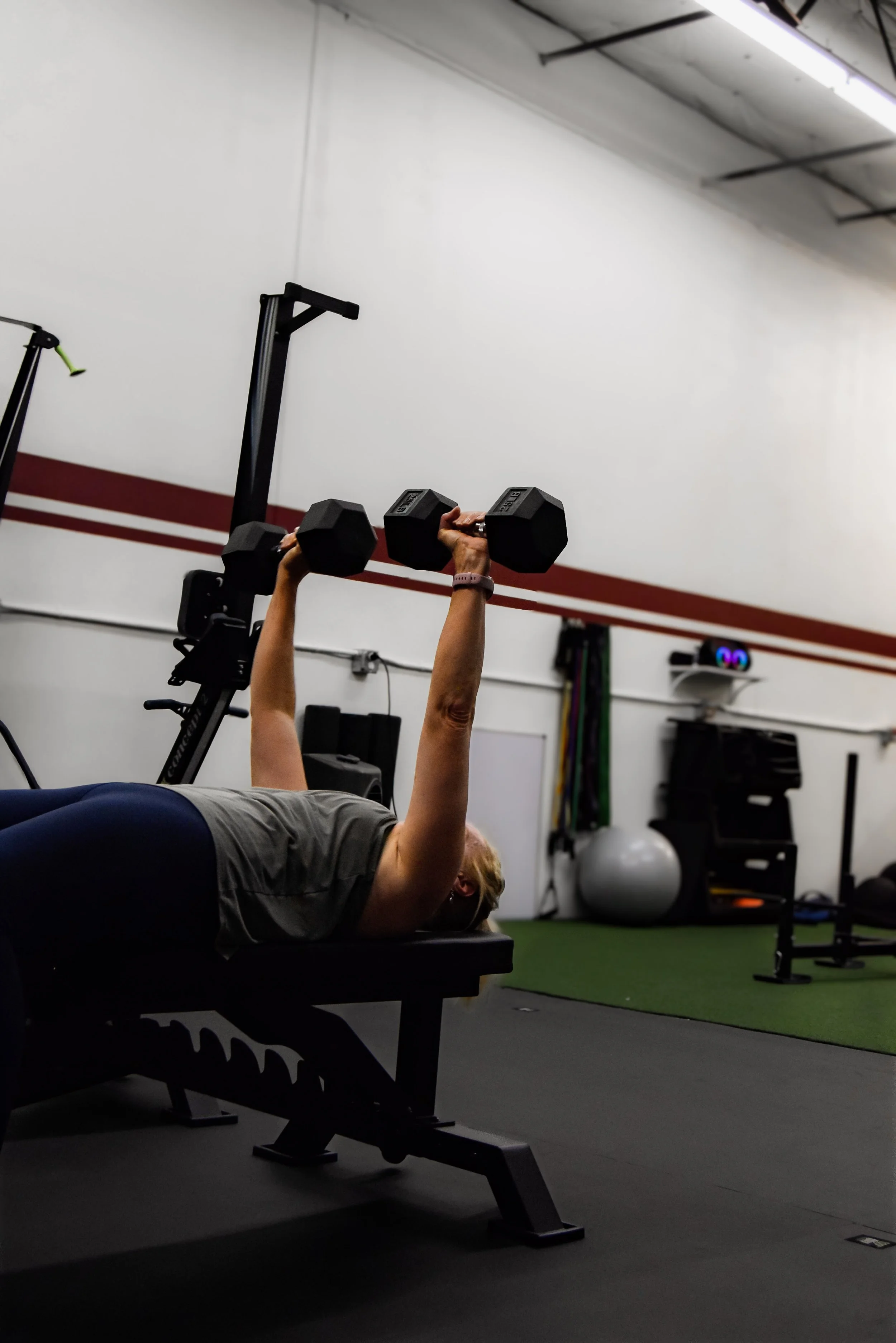 A person lying on a bench in a gym lifting a pair of black dumbbells above their chest, with gym equipment and a white wall in the background.