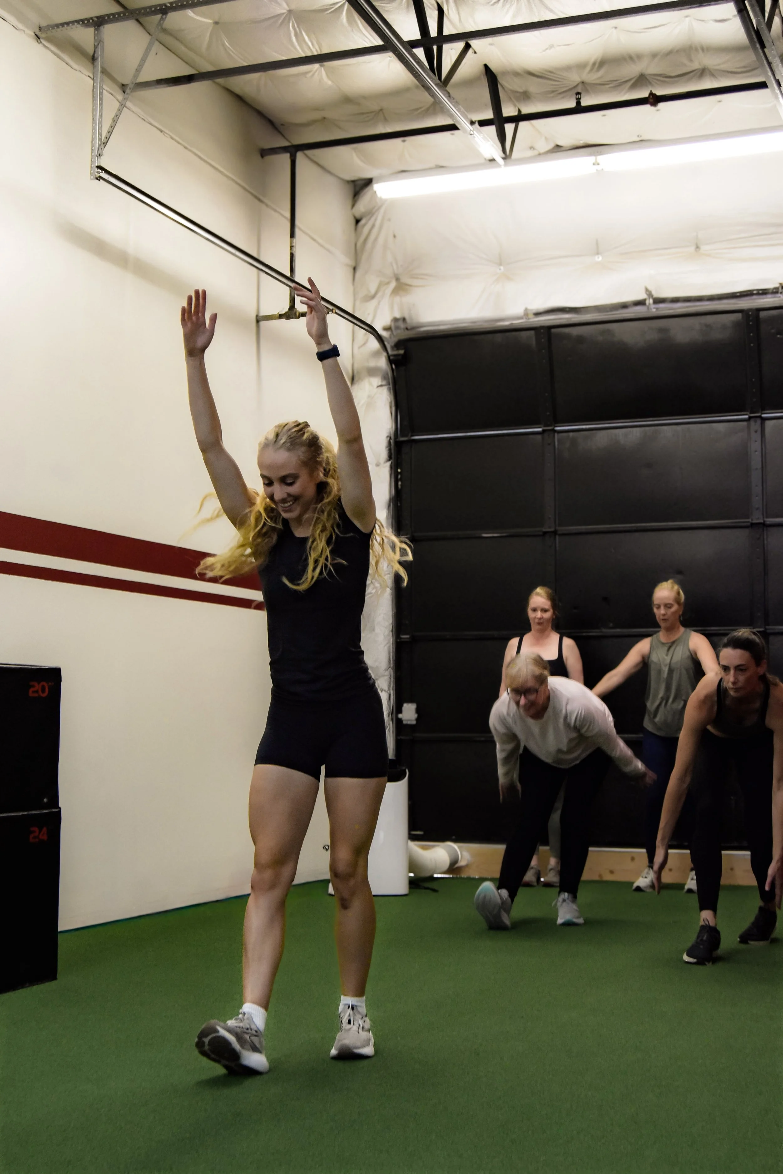 A woman with long, curly blonde hair is smiling and exercising in a gym, wearing a black T-shirt, black shorts, and gray sneakers. She has her arms raised above her head, and a group of people behind her are participating in a workout class, performing leg stretches on a green mat. The gym has a white wall with red stripes, black lockers or containers, and a high ceiling with exposed insulation and metal support beams.