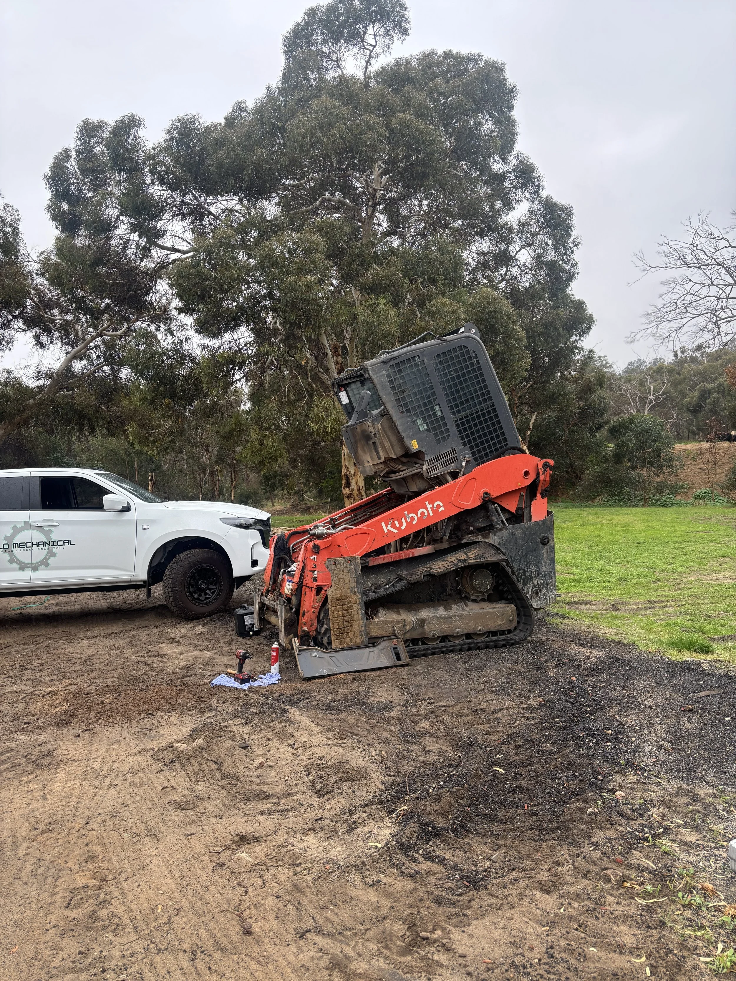 A construction site with a white vehicle on the left and a small orange excavator on the right. The excavator's bucket is raised, and there are tools and a piece of paper on the ground in front of it. Large trees and a grassy area are in the background, and the ground is a mix of dirt and grass.