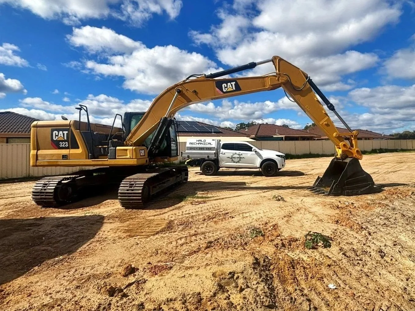A yellow Caterpillar excavator with tracks on sandy ground, with a white pickup truck and a row of houses in the background under a partly cloudy sky.