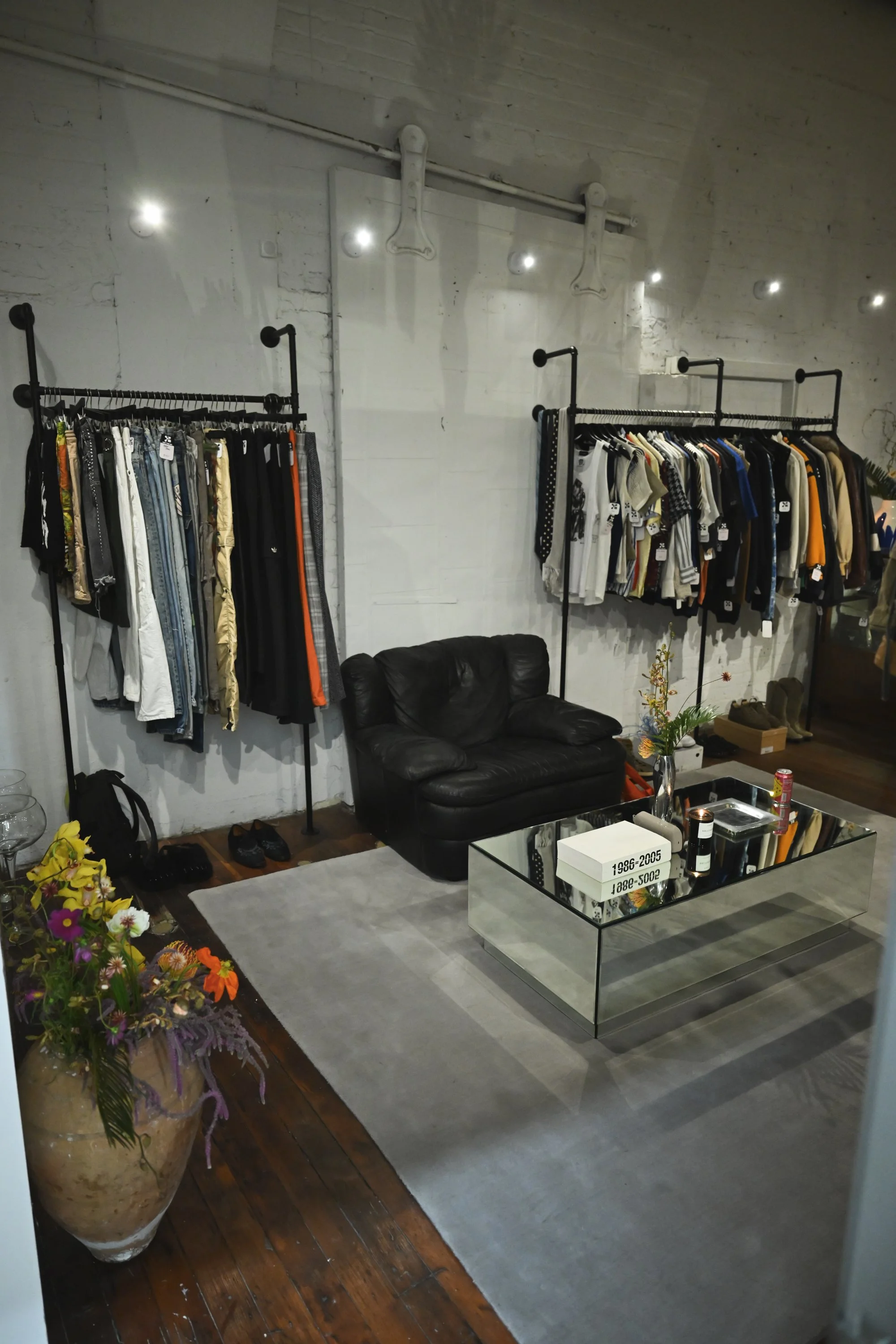 Interior of a clothing store with two rackings of clothing, a black leather couch, a coffee table, and a large floral arrangement in a tall terracotta pot. The store has a white brick wall, exposed piping, and decorative bone-shaped hooks.