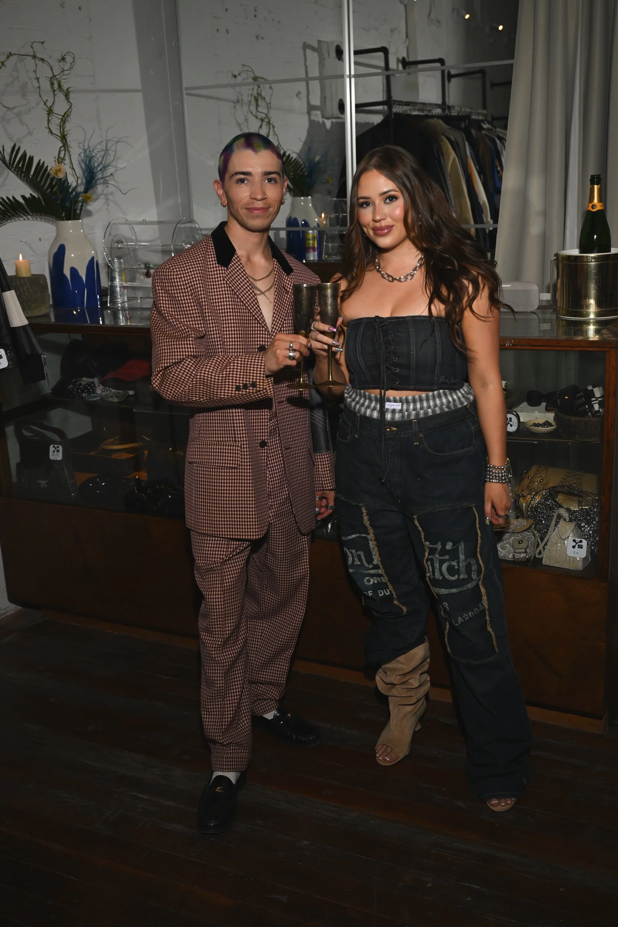 Two people standing indoors, holding champagne glasses and posing for a photo. The person on the left has rainbow-colored hair and is wearing a checkered suit. The person on the right has long brown hair and is wearing a black crop top, distressed jeans, and beige boots. Behind them is a clothing rack, a mirror, and decorative plants.