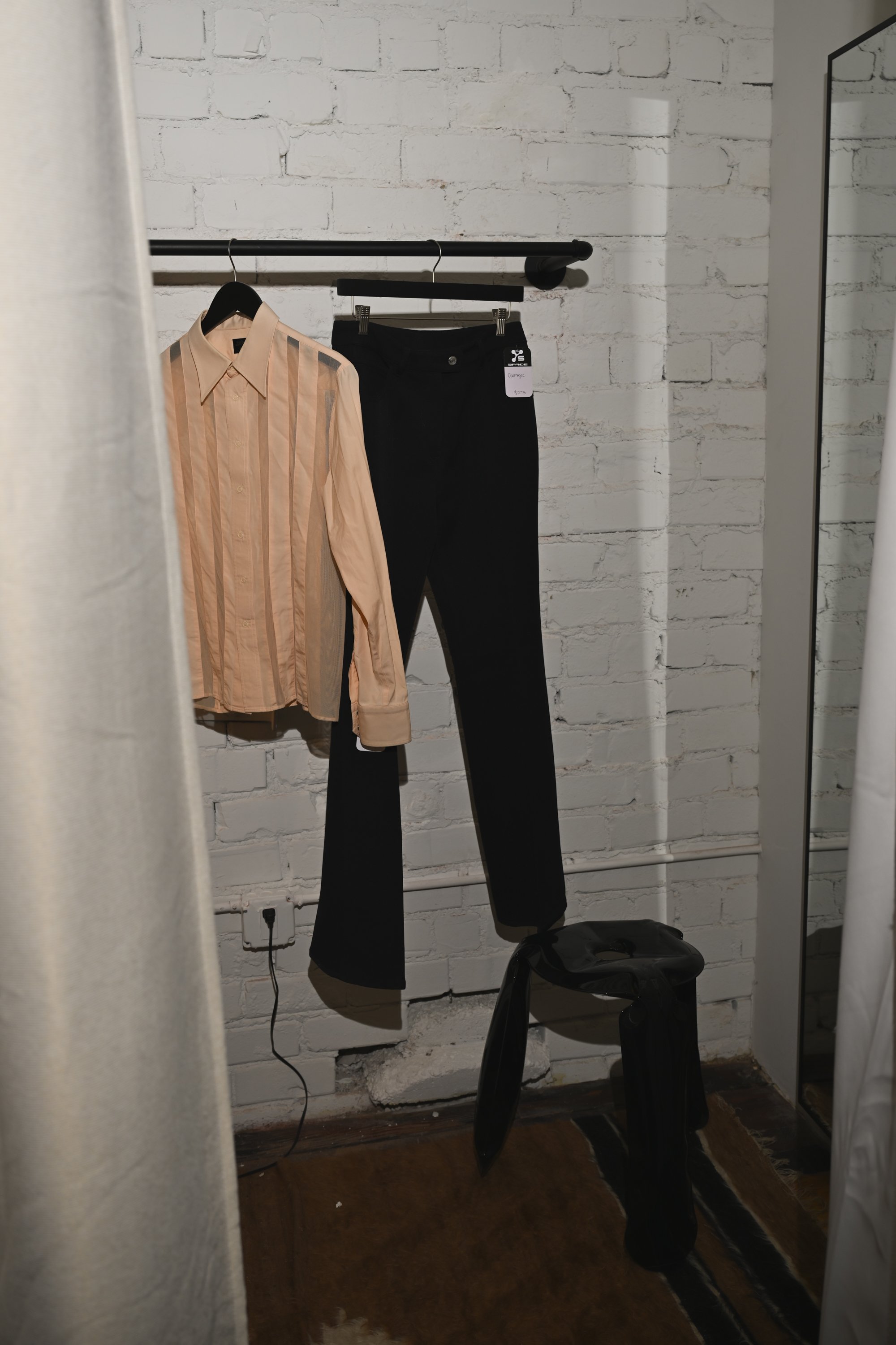 A clothing rack with a beige striped shirt and a pair of black pants hanging in front of a white brick wall, with a mirror reflecting part of the room and a black chair on the floor.
