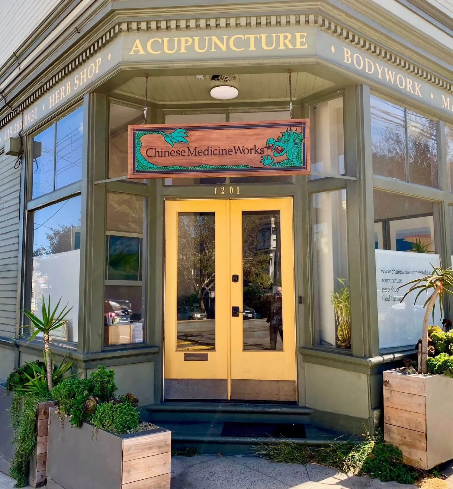 Front exterior of a storefront with signs for acupuncture, herbal shop, and bodywork, featuring large glass windows, a yellow door, and potted plants outside.