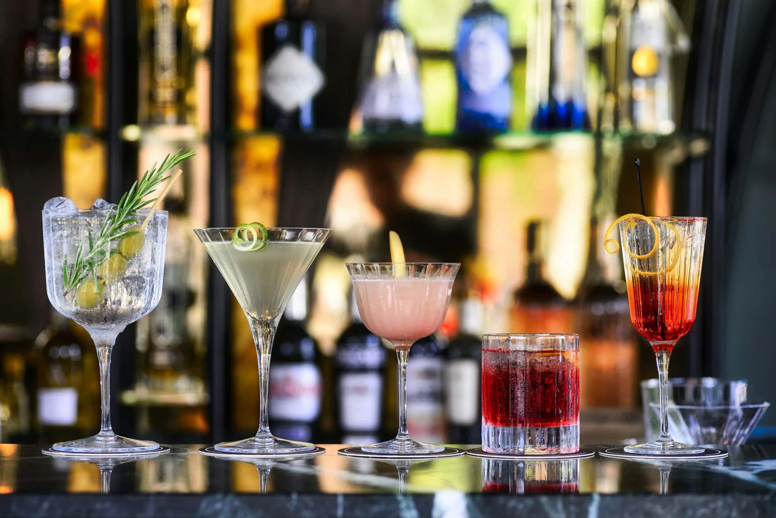 Various colorful cocktail beverages in elegant glassware on a bar counter with a blurred background of liquor bottles.