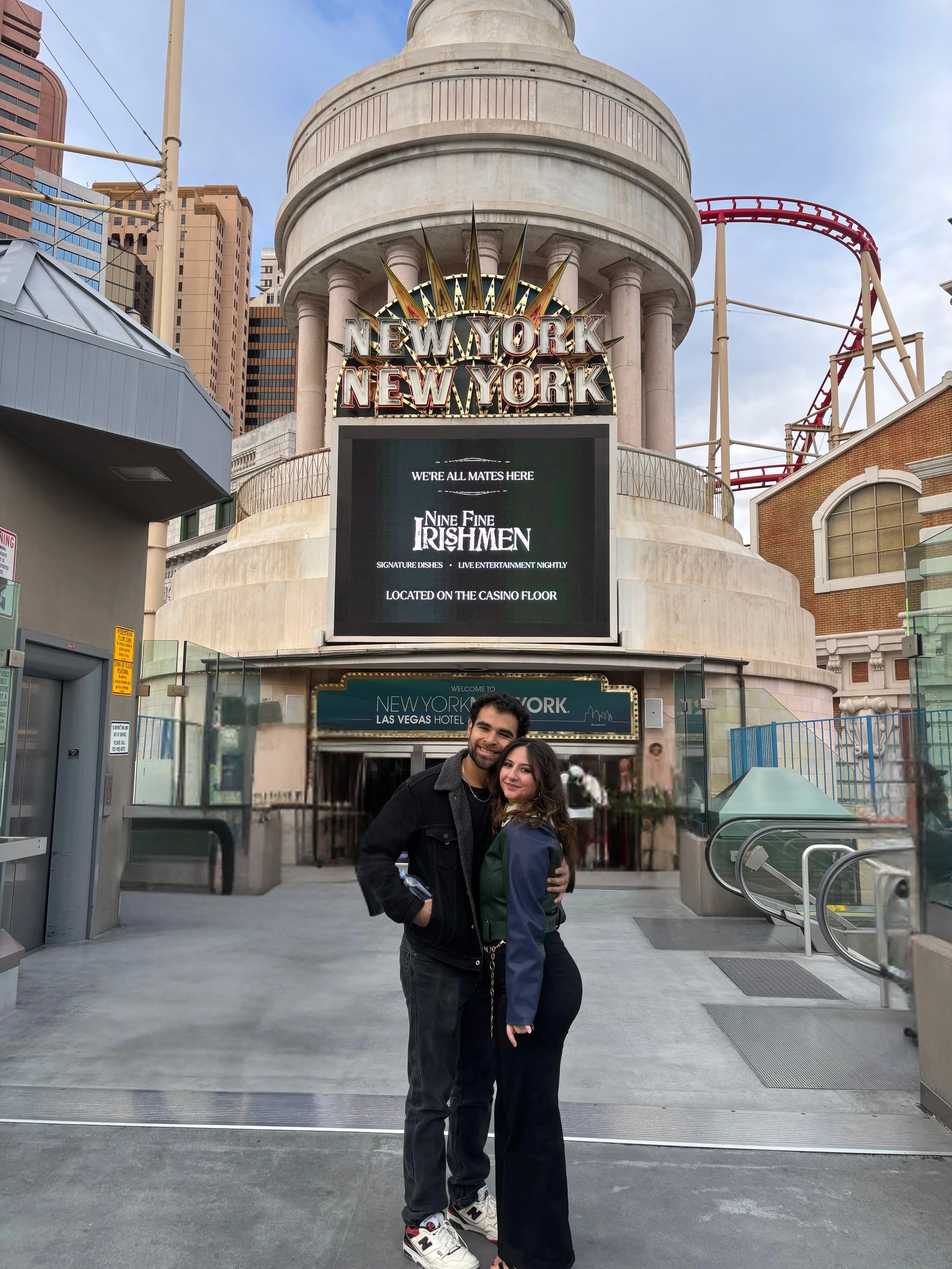 A smiling man and woman stand close together, arms around each other, in front of the New York-New York Hotel and Casino entrance in Las Vegas. The hotel features a replica of the New York City skyline and a roller coaster track in the background.