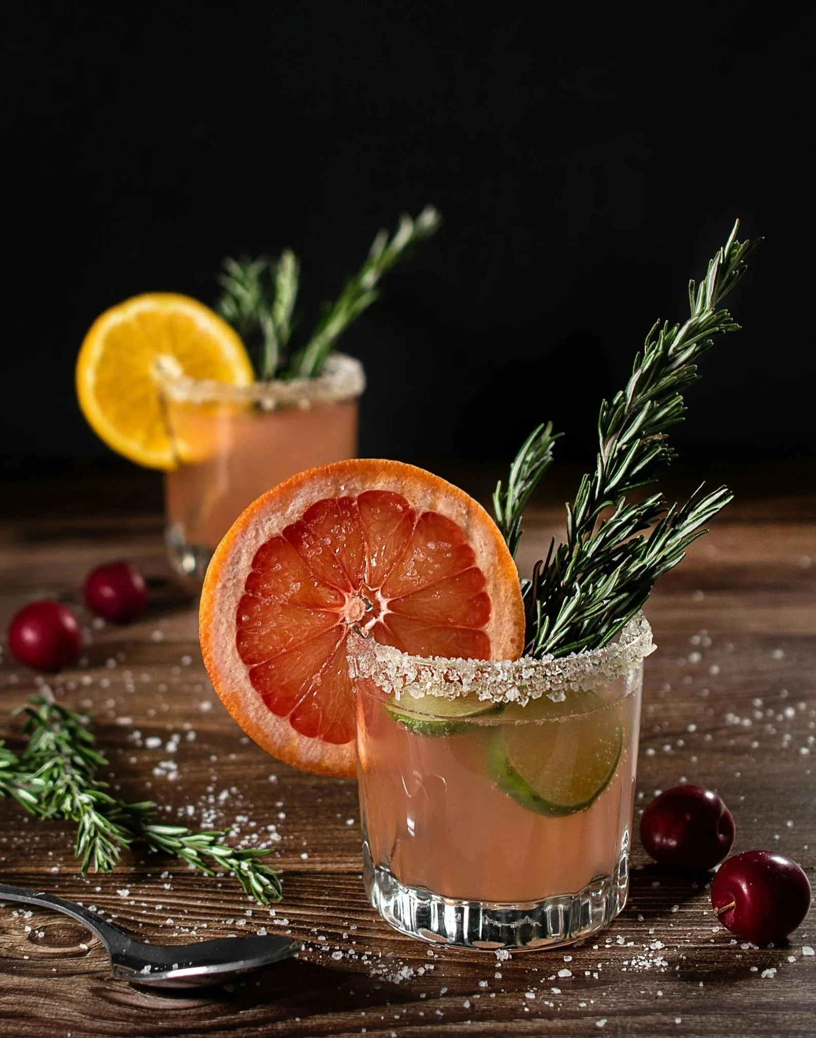 A close-up of two cocktails with salted rims, garnished with slices of grapefruit, lemon, cucumber, and sprigs of rosemary, on a wooden table with scattered spices and cherries.