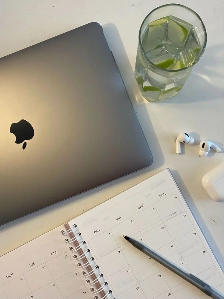 A silver MacBook, a glass of water with lime wedges, wireless earbuds, a white wireless charging case, a silver pen, and an open planner on a white desk.