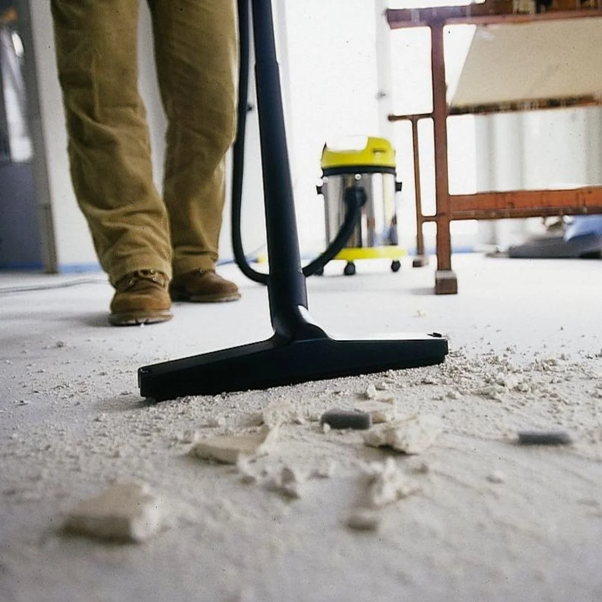 Close-up of a person using a floor sander on a dusty concrete surface in a room under renovation; the person's legs and boots are visible, with workshop tools and furniture in the background.