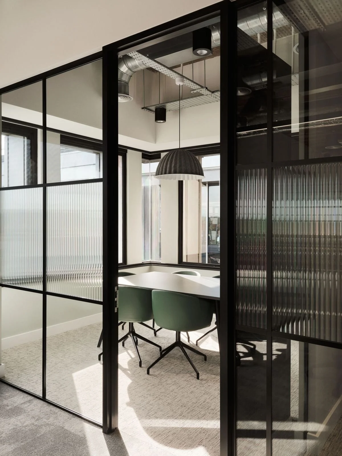 Modern office conference room with glass walls, a white table, and three green chairs, illuminated by natural light from large windows, with a black pendant lamp hanging overhead.