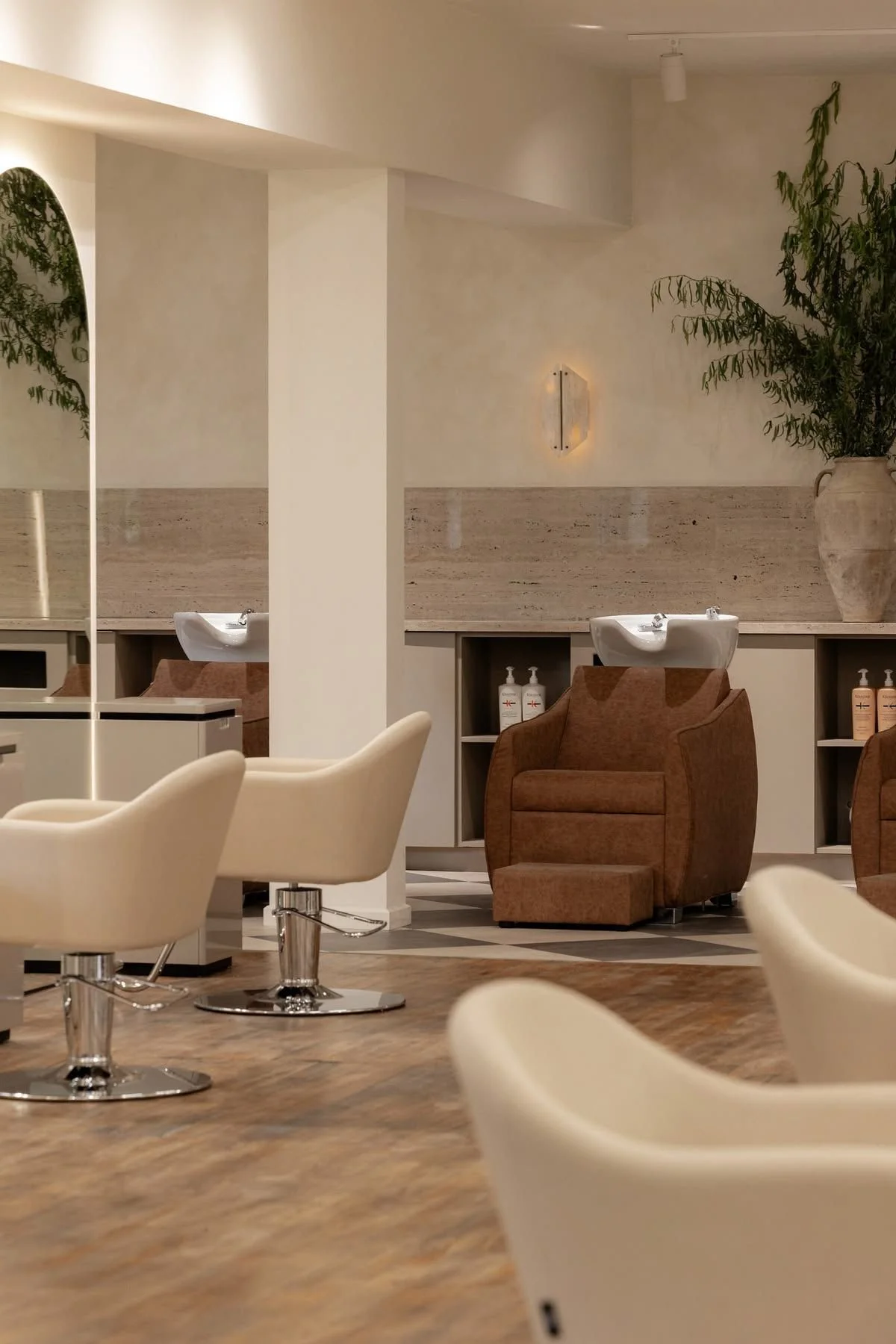 Interior of a modern hair salon with washing stations, white styling chairs, brown reclined chairs, and green plants.