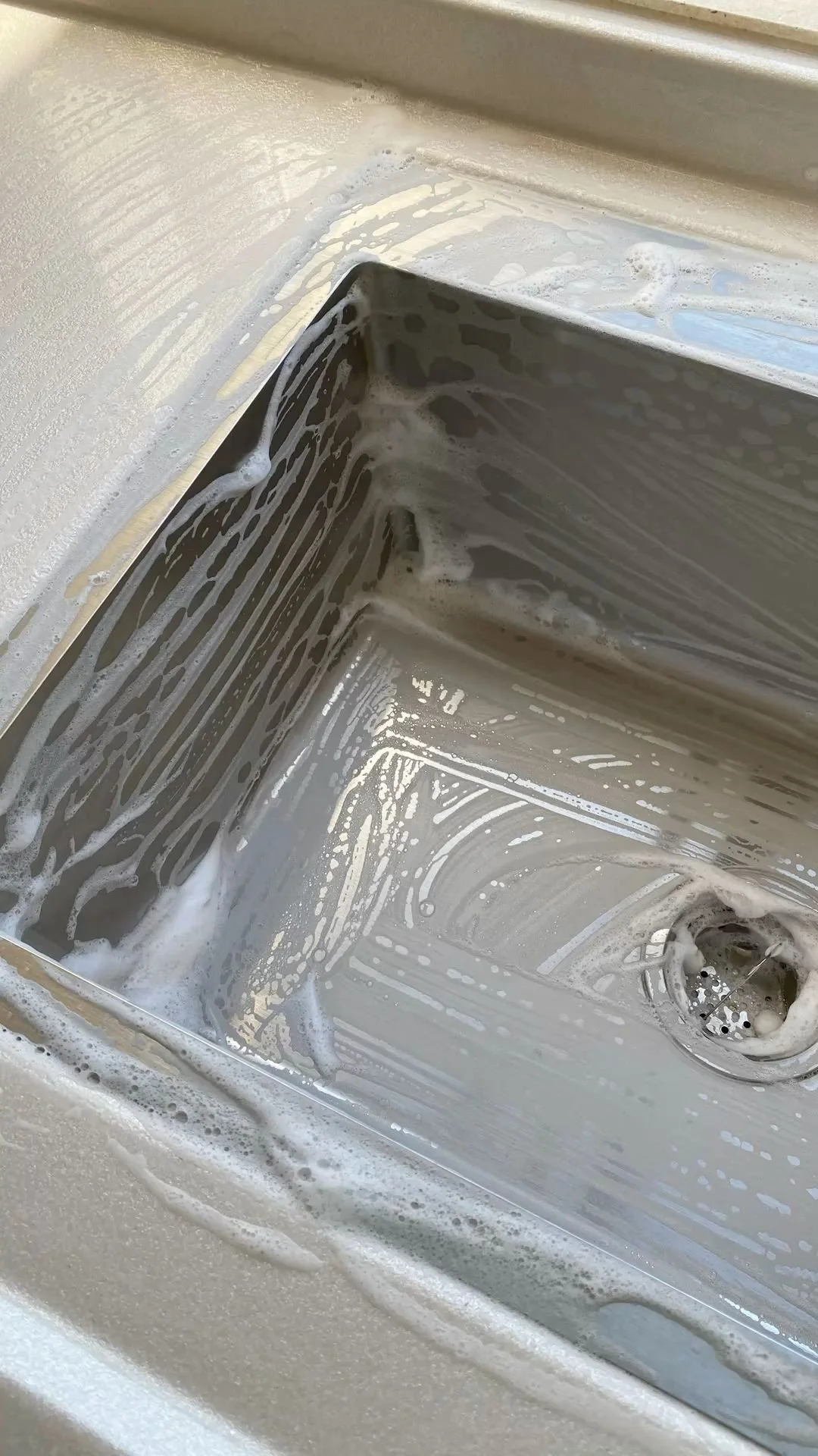 Inside a stainless steel kitchen sink with soap suds and water residue.