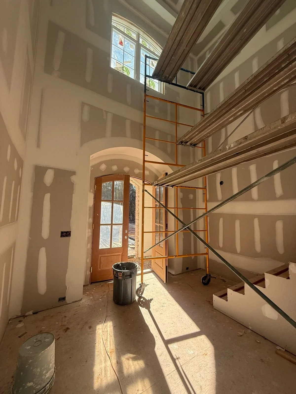 Interior of a house under construction with drywall walls, a door, and a large window. Construction scaffolding and metal supports are present inside, with sunlight coming through the window and door.
