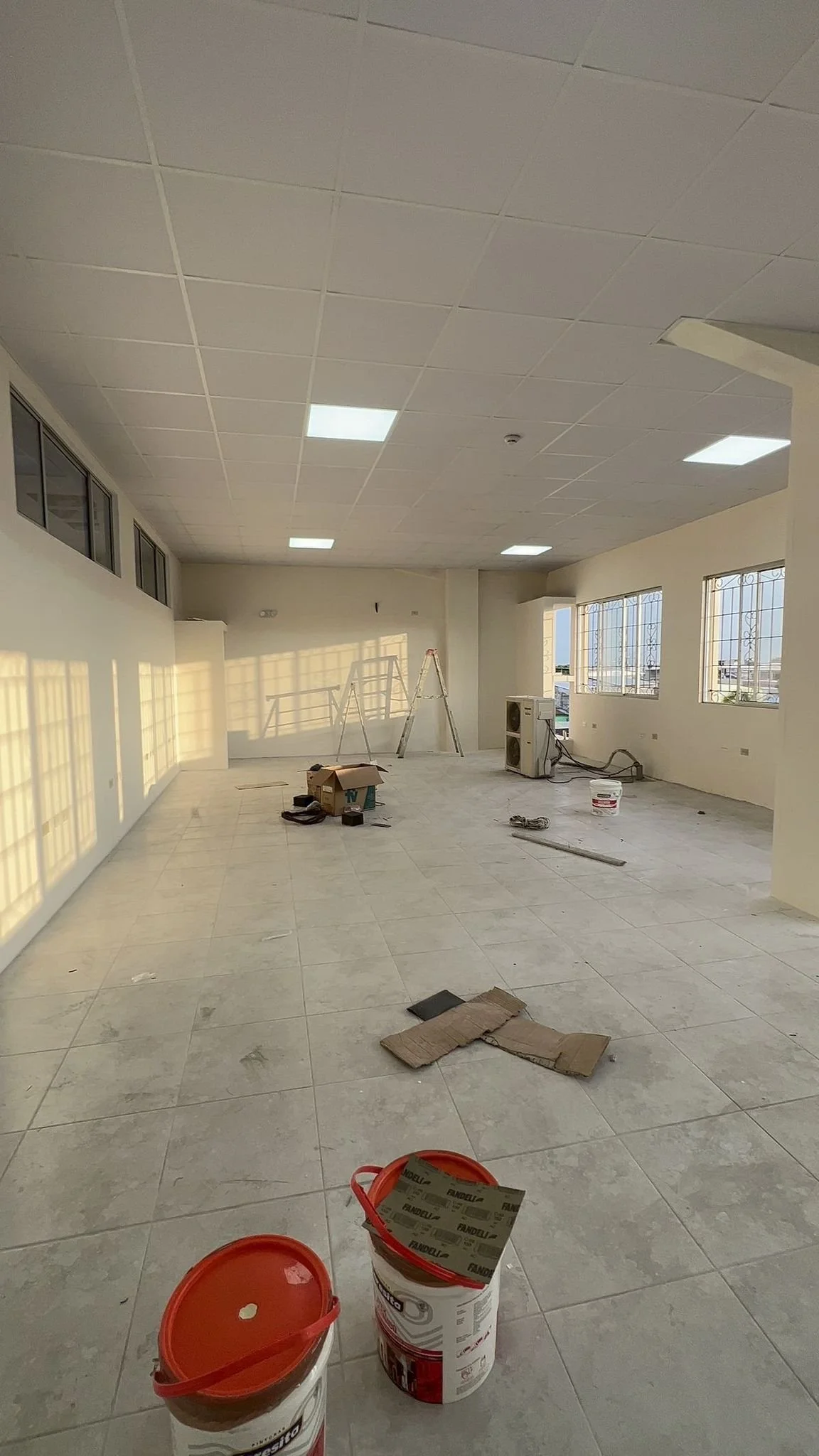 Empty room under construction with tile flooring, ceiling tiles, and several windows, construction tools, ladders, and buckets of paint scattered around.