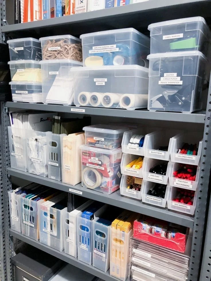 Shelf of organized office supplies, including labeled plastic containers with rubber bands, tape rolls, scissors, pens, markers, and various other stationery items.