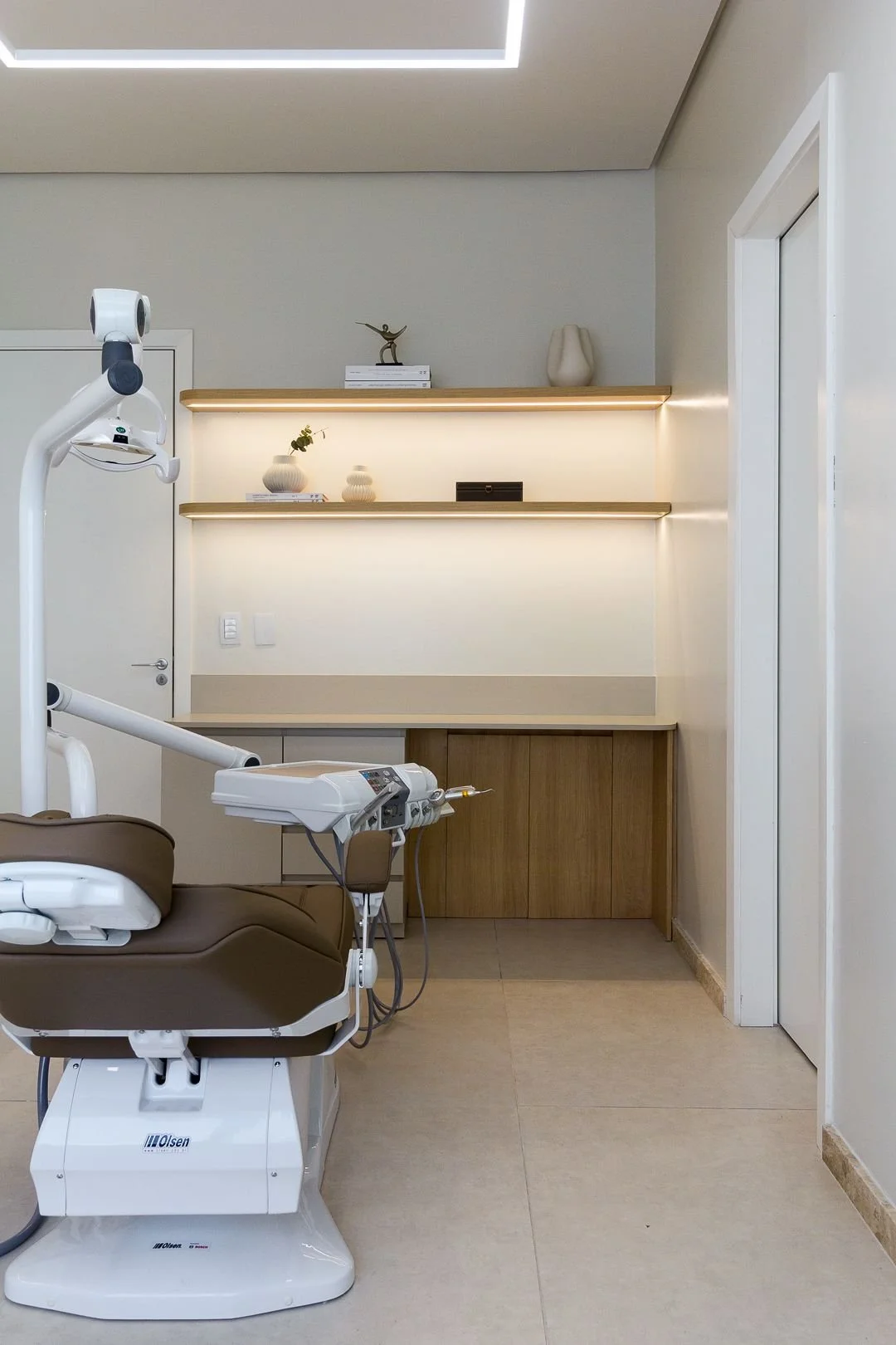 Dental examination room with dental chair and equipment, shelves with decorative vases and books, and a light-colored wall.
