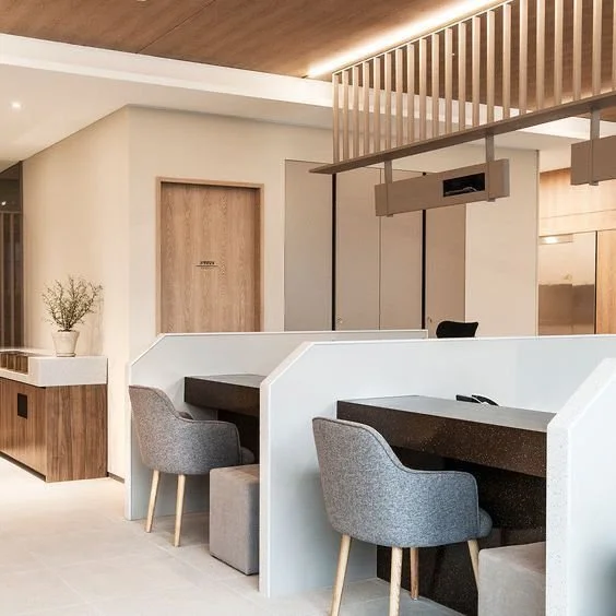 Modern office workspace with two workstations separated by a low white wall, each with a black desk and gray upholstered chair, and a wooden ceiling and wall accents.
