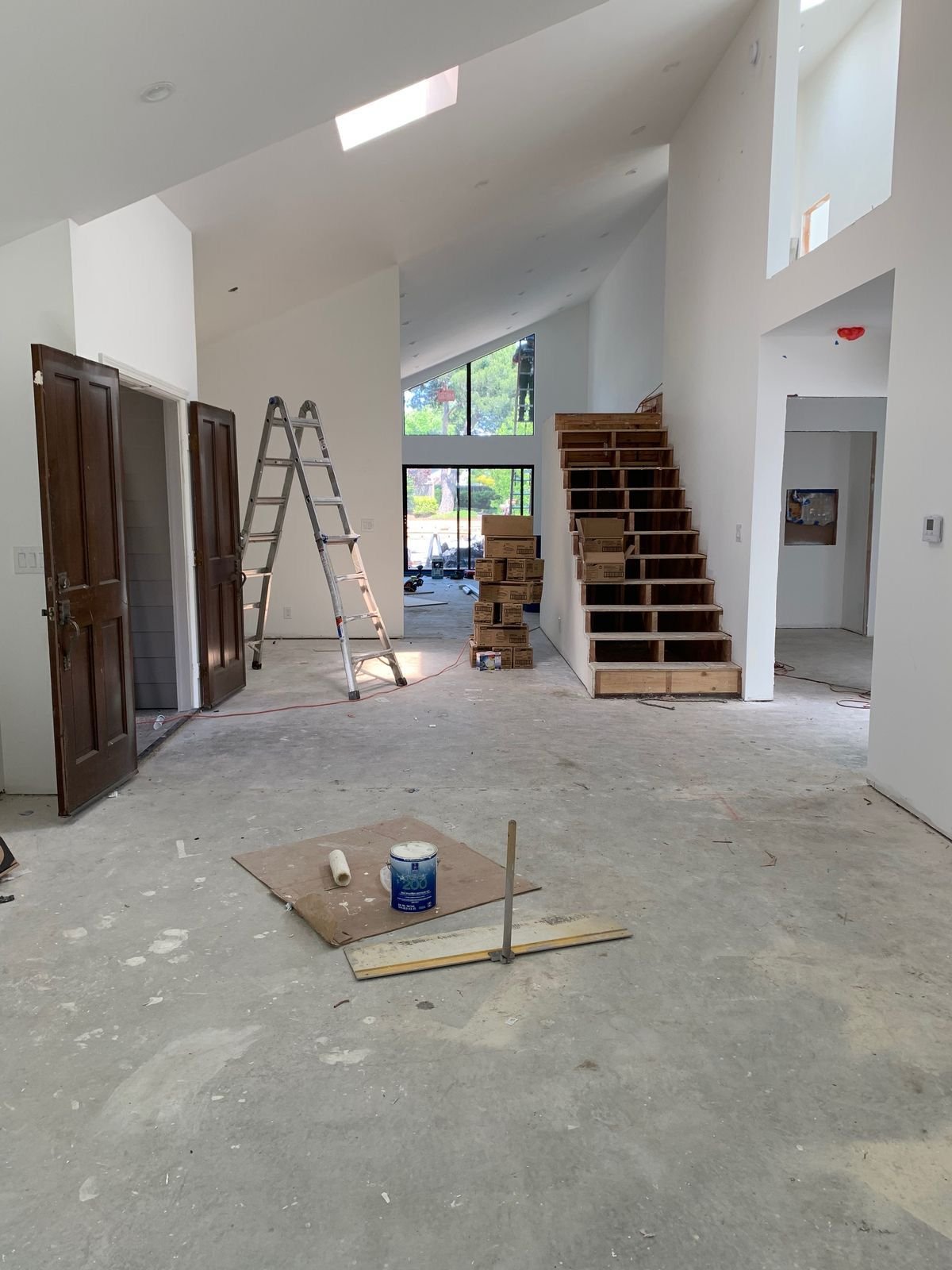 Interior of a house under construction with an unfinished staircase, open doorways, a ladder, construction materials, and tools scattered on the floor.