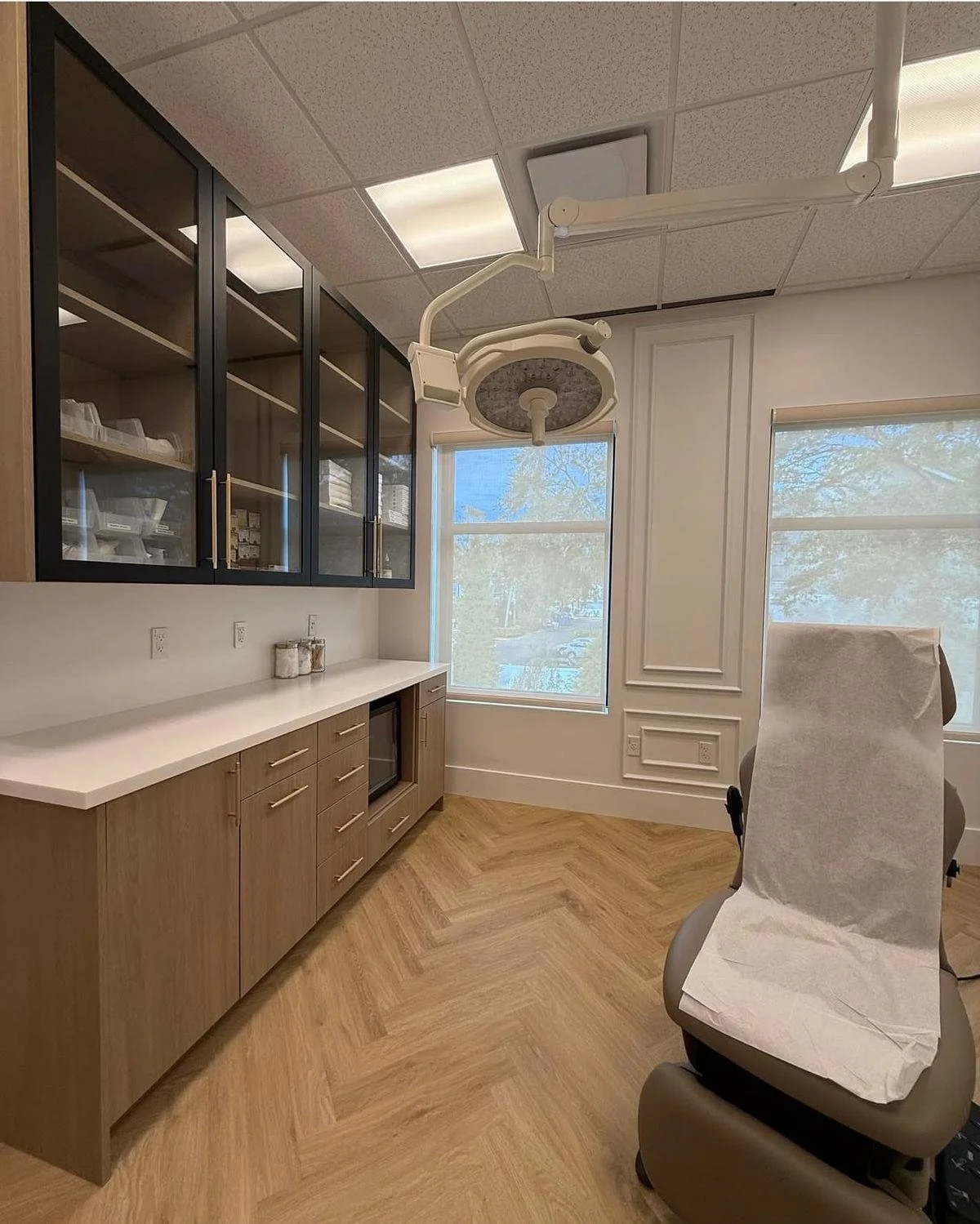 A clean, modern dental office with a dental chair covered in white paper, wooden cabinets, a dental light overhead, and large windows allowing natural light.