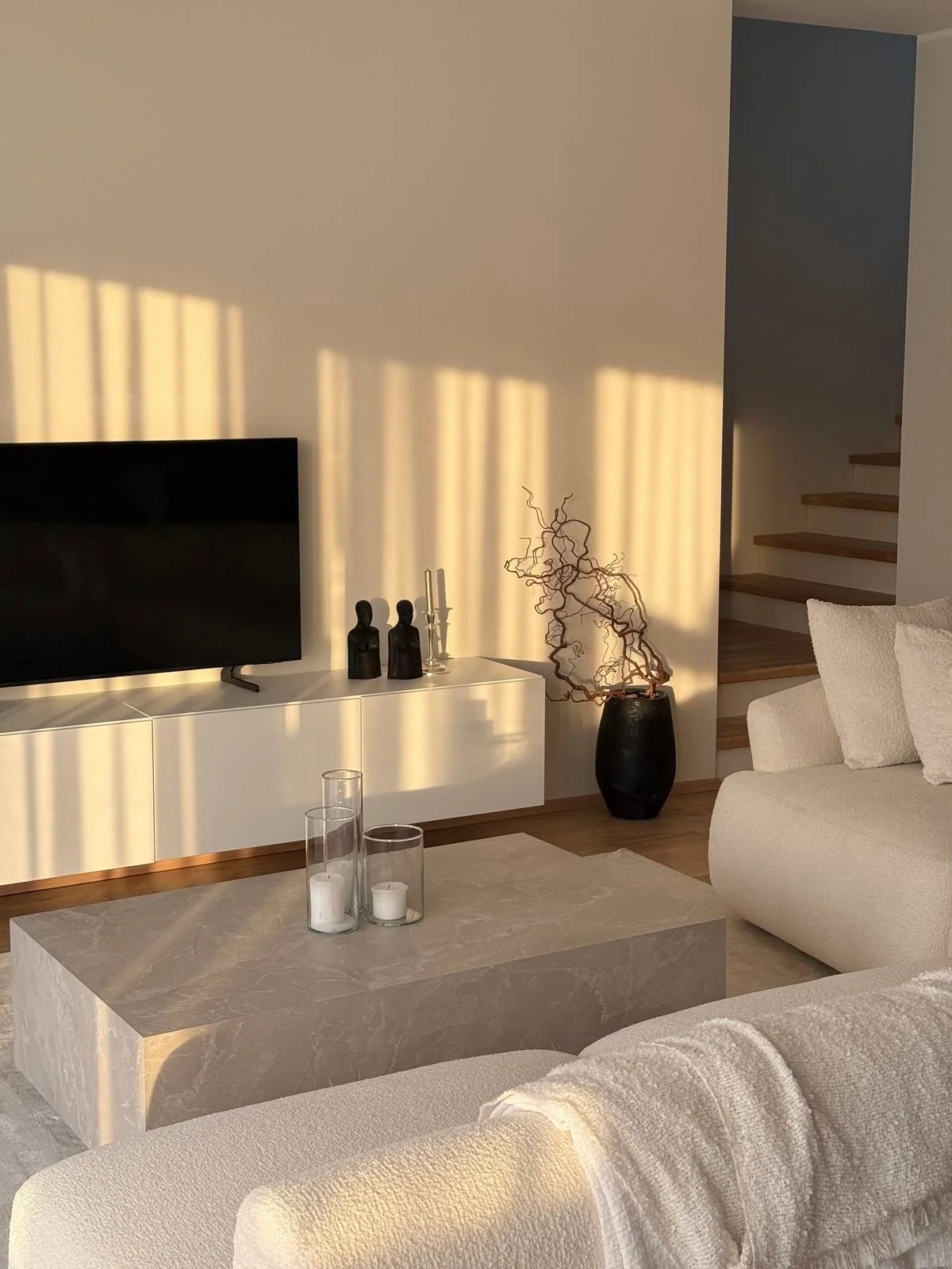 Living room with a black TV on a white console, black decorative sculptures, candles in glass holders, a black vase with twisted branches, a cream sofa, a marble coffee table, wooden stairs, and sunlight casting shadows on the wall.