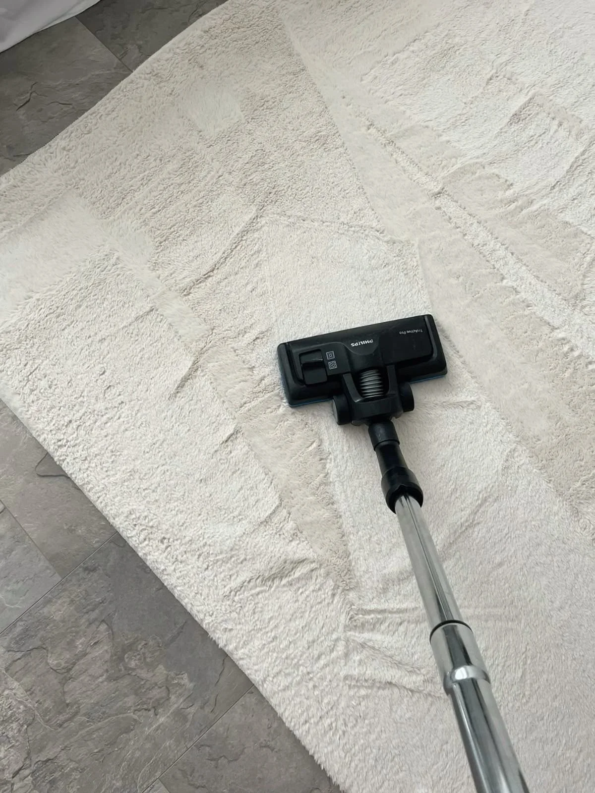 A vacuum cleaner head is on a cream-colored, textured rug, with a metal extension tube attached, in a room with gray tile flooring.