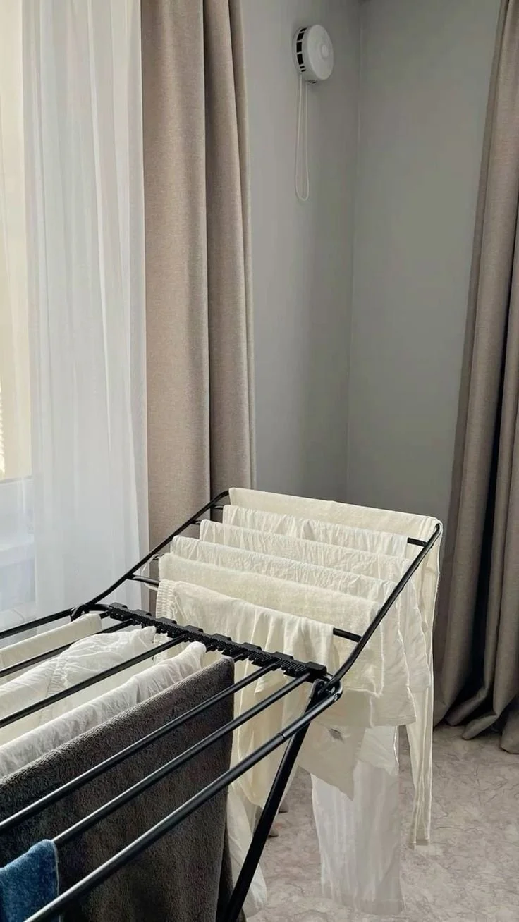 Laundry drying rack with white and dark-colored clothes inside a room with beige curtains and a smoke detector on the wall.