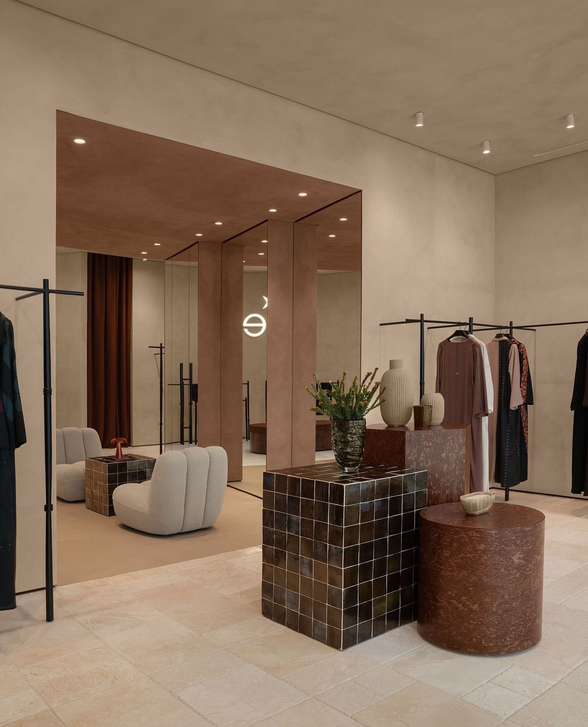 Elegant clothing boutique interior with beige walls, modern furniture, and clothing racks displaying dresses and garments. Decor includes vases, a plant, and textured furniture pieces, creating a stylish and minimalist ambiance.