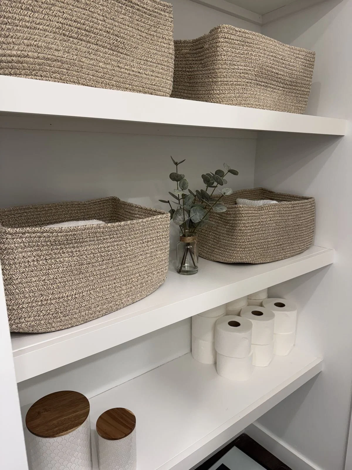 White shelving unit with woven baskets, a small glass vase with greenery, and rolled toilet paper.