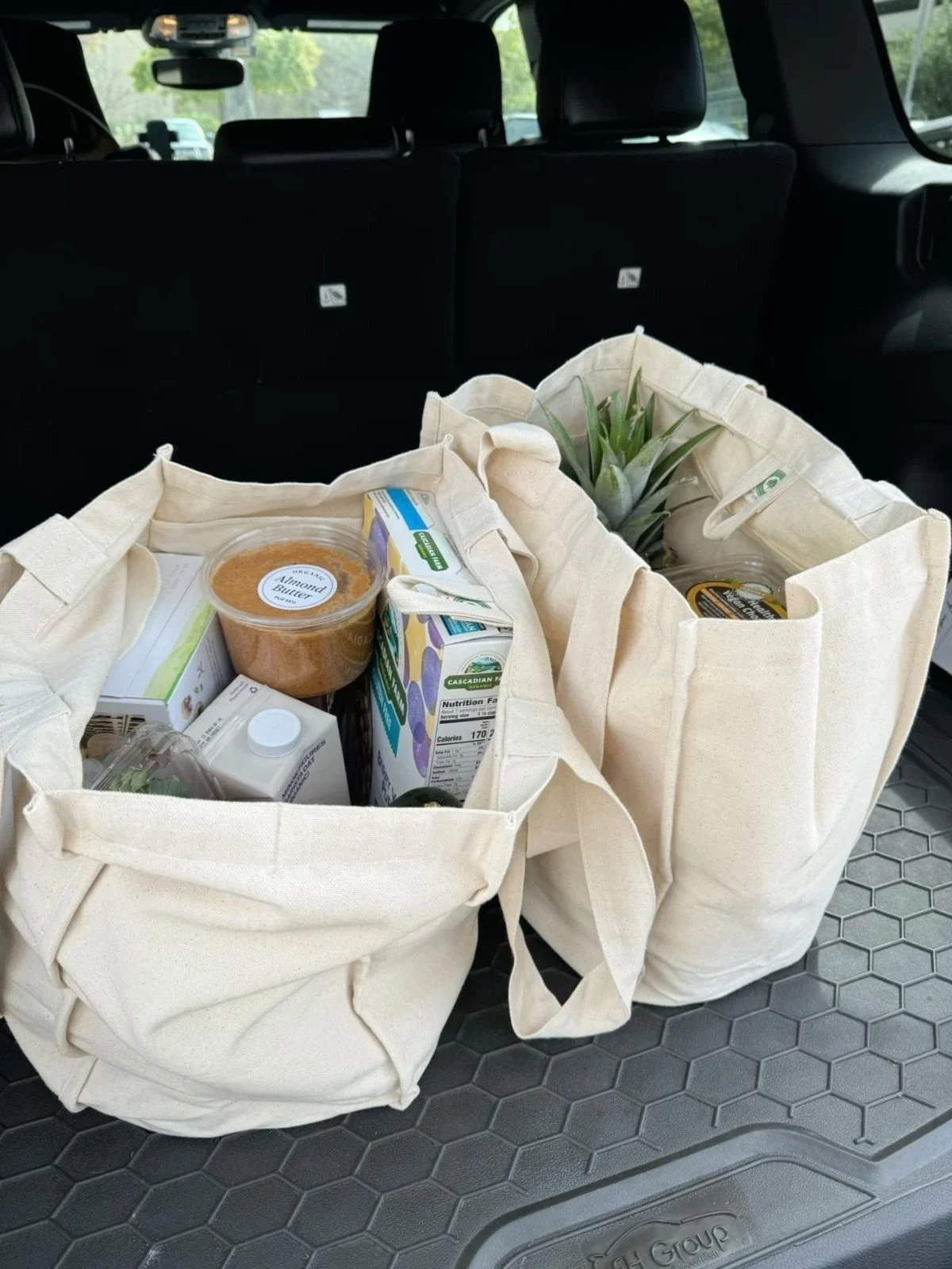 Two canvas tote bags filled with groceries, including pineapple, almond butter, milk cartons, and other food items, placed on the trunk floor of a vehicle.