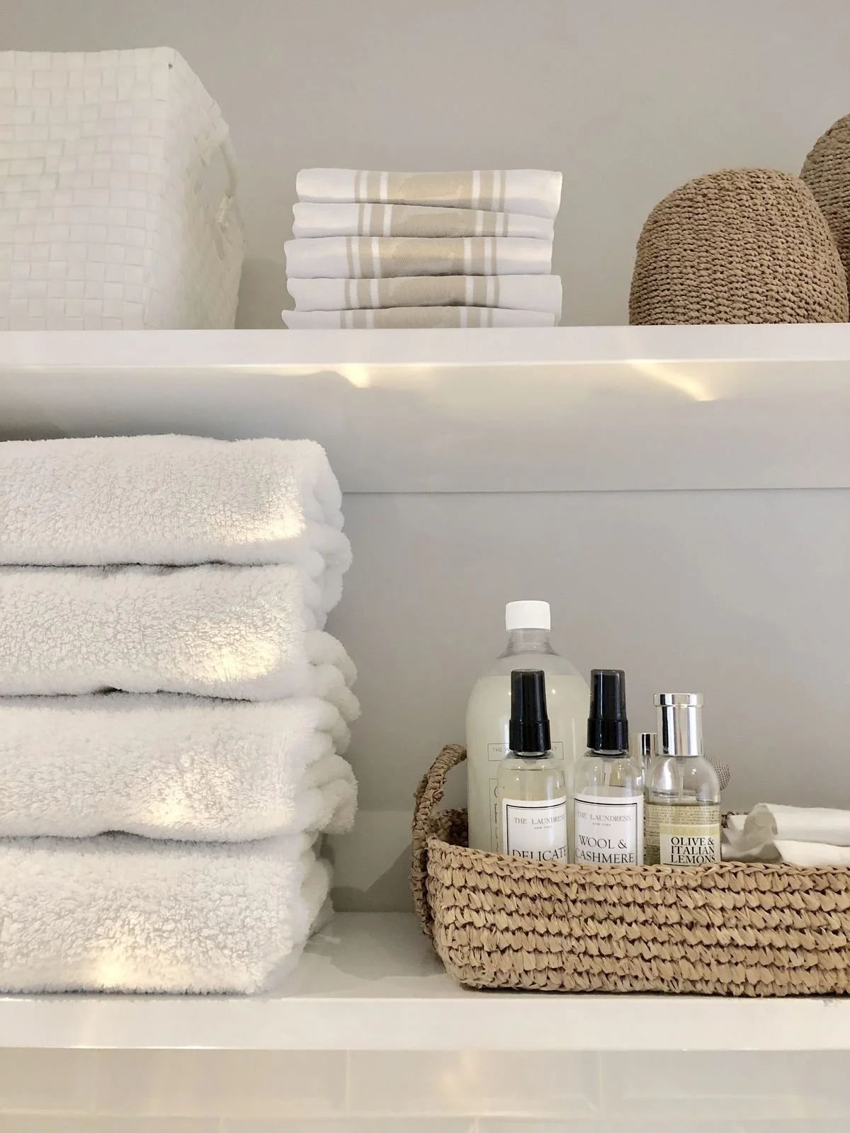 Stacked white towels, folded beige-striped towels, round brown textured objects, and a basket with linen spray bottles on white shelves.
