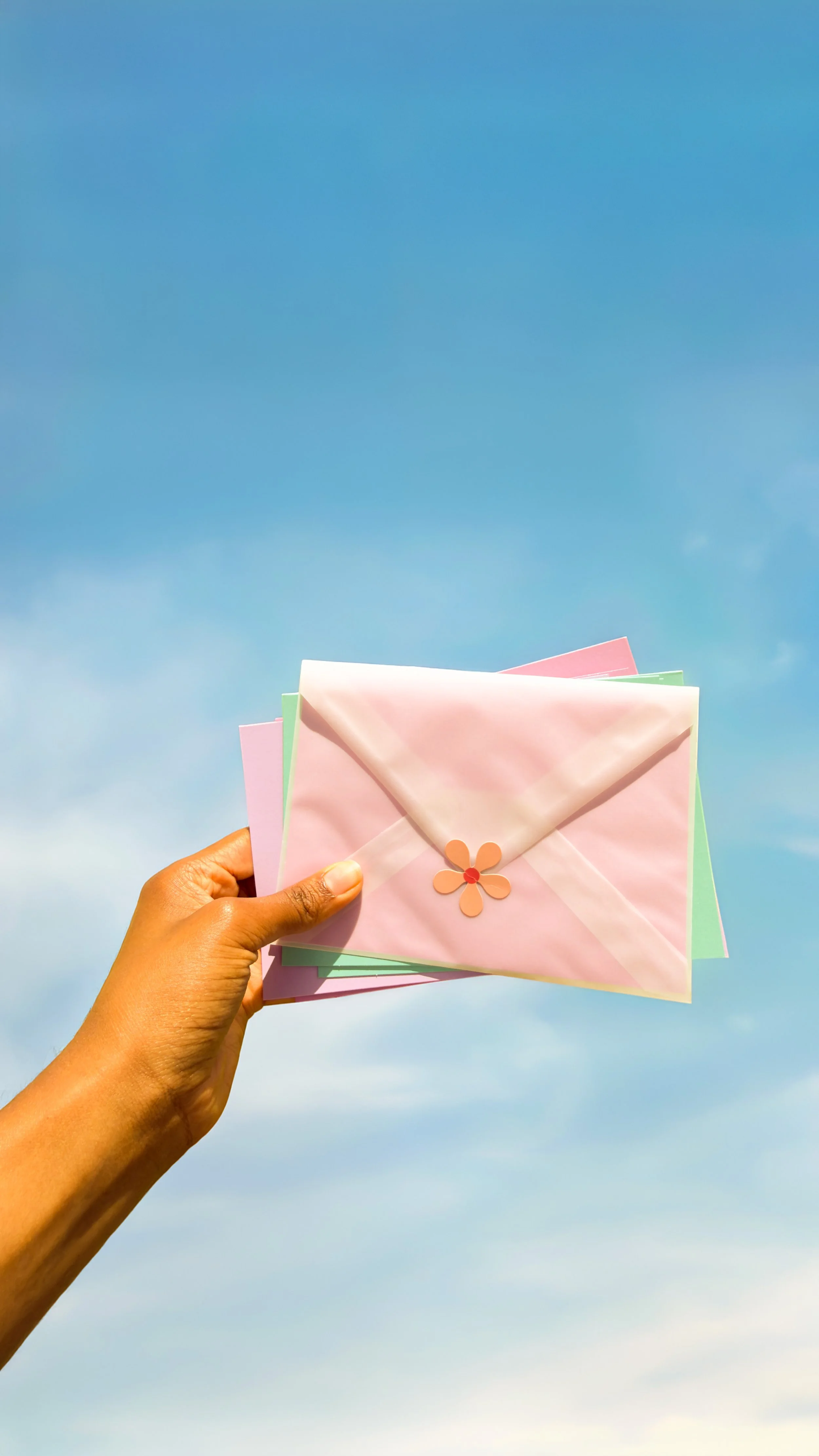 Person holding pastel-colored envelopes with a pink flower decoration against a blue sky background.