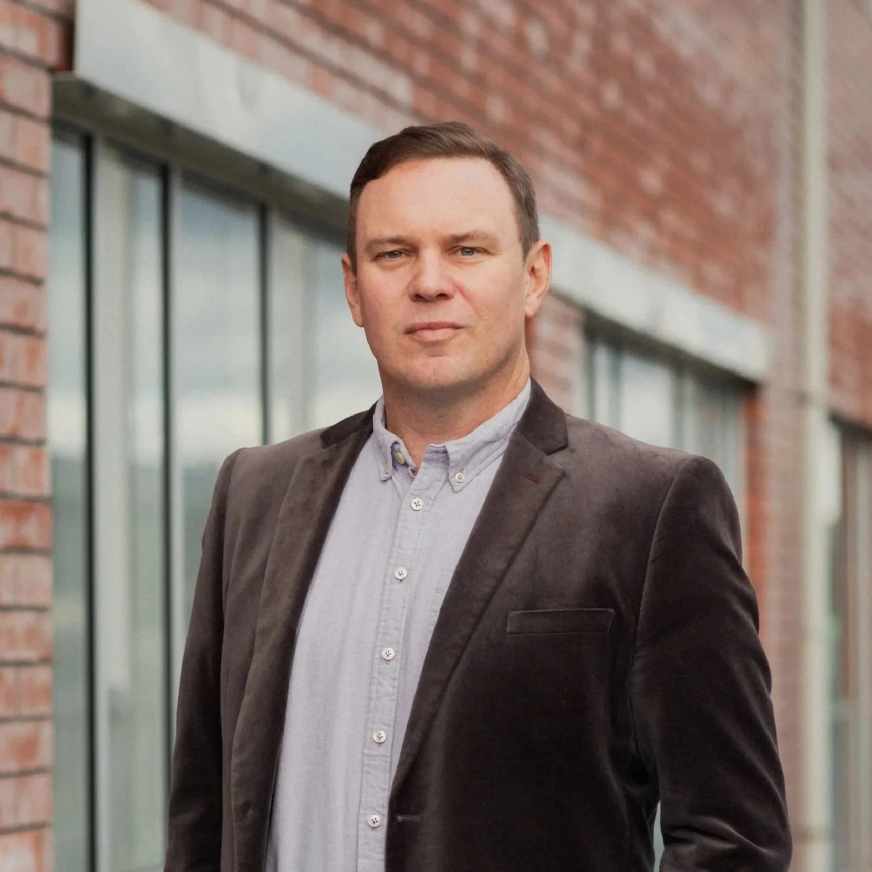 It is Sam Houliston in a grey blazer and light gray button-up shirt standing outdoors near a brick building with glass windows. He is probably thinking about employment law or death metal.