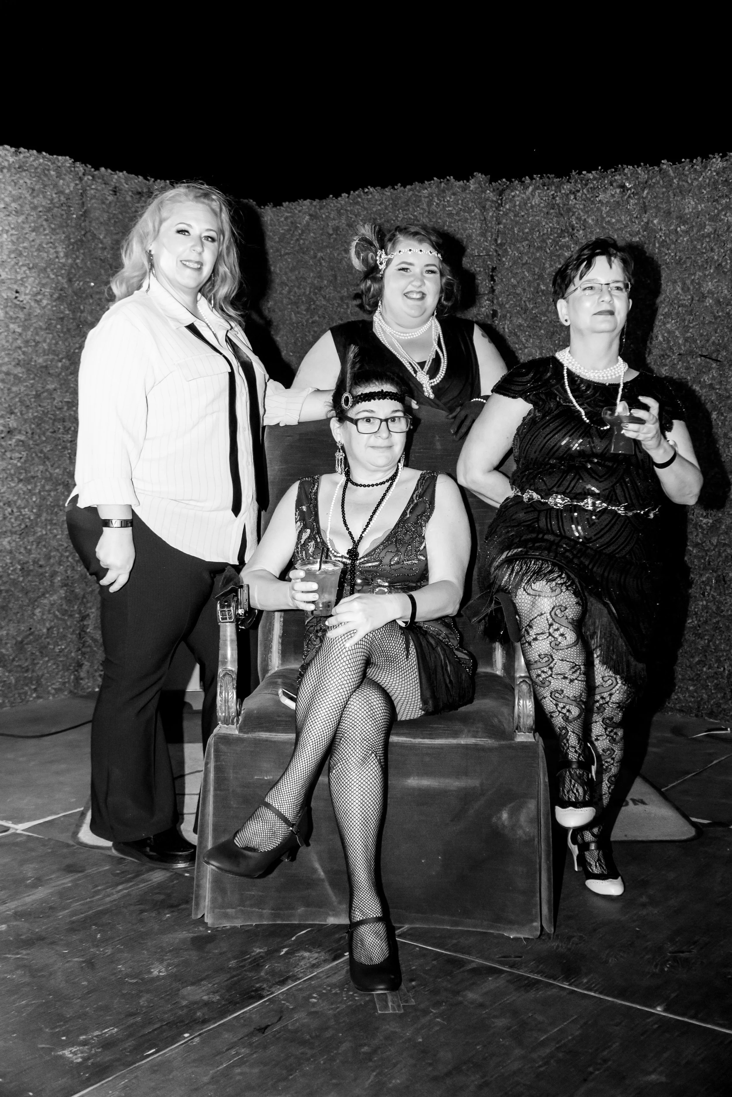 Four women dressed in vintage 1920s costumes posing together on a stage against a textured wall. They are wearing flapper dresses, beads, headbands, and fishnet stockings, with one holding a drink and seated on a chair, and the others standing around